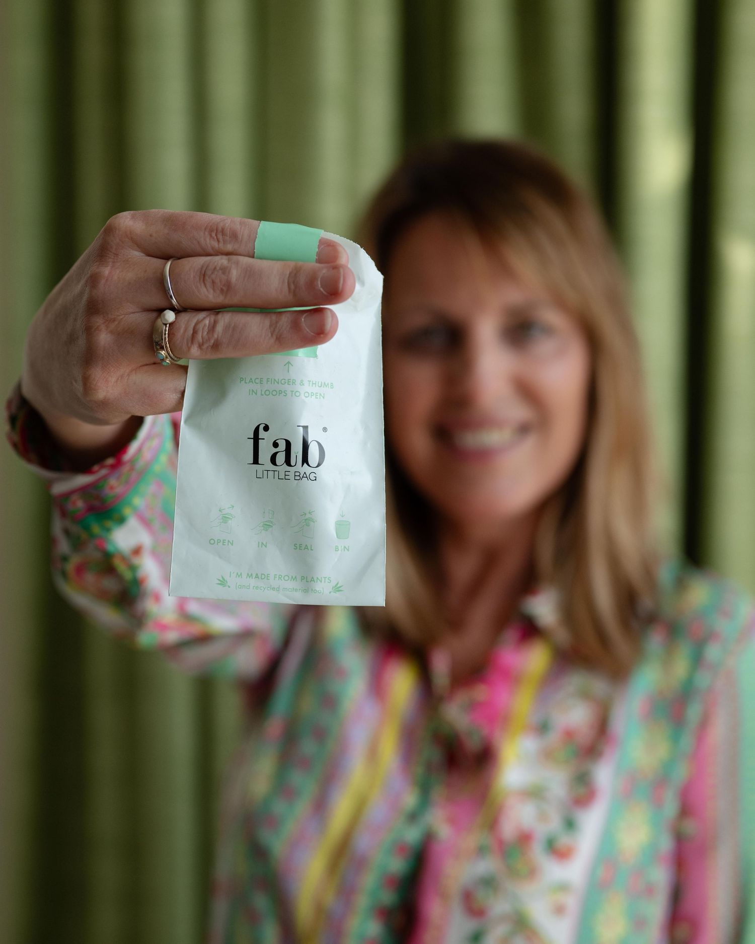 Woman in colorful floral blouse holding Fab Little Bag close to camera with green curtain backdrop.