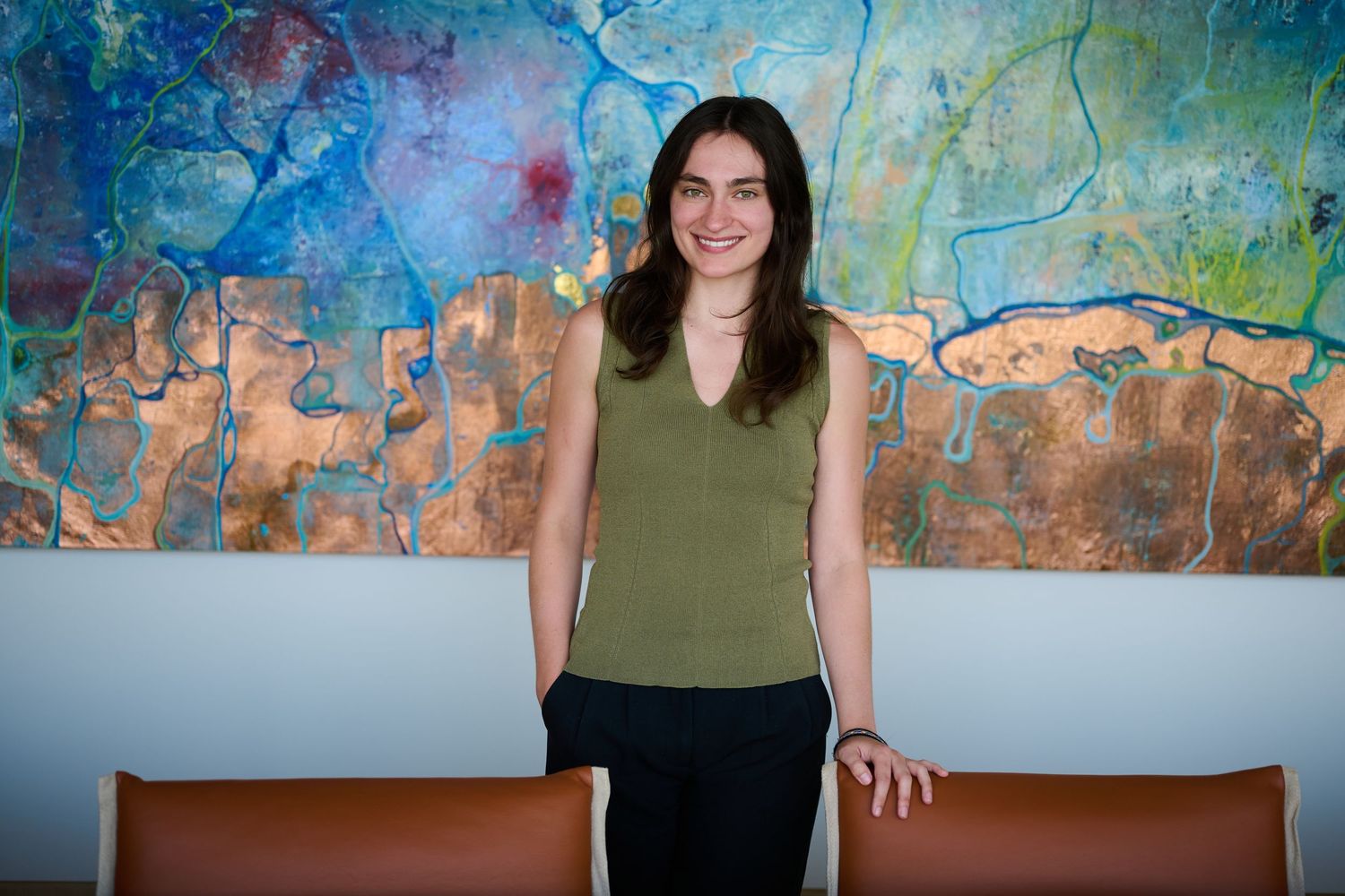 Young woman in olive top smiling before colorful abstract painting in modern art gallery setting.