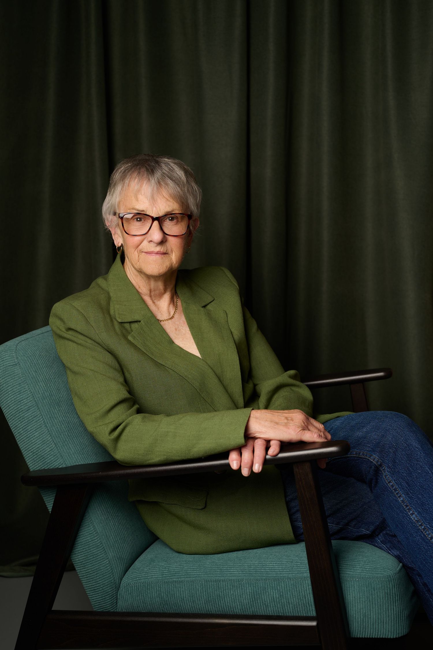 Elderly woman with glasses in green blazer and jeans seated in a teal chair against dark green curtains.