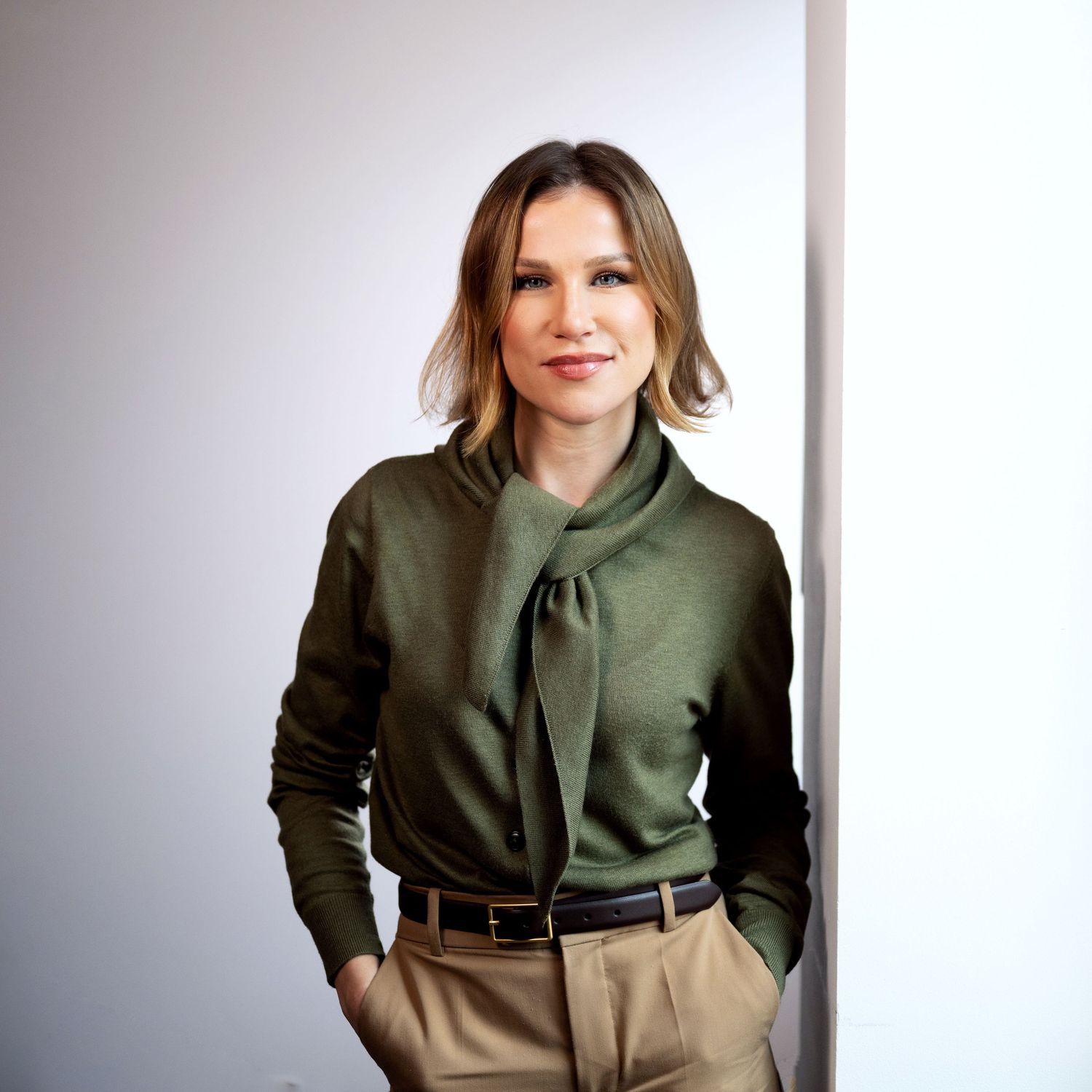 Woman in olive green tie-neck blouse and tan trousers, hands in pockets, leaning against white wall.