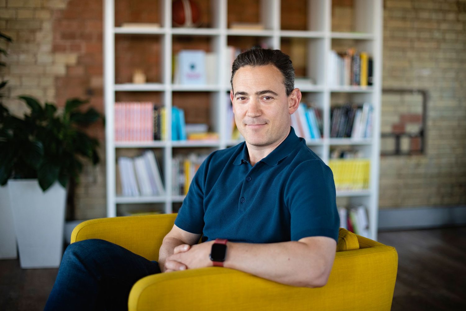 Smiling man in navy polo seated on yellow chair in modern office.
