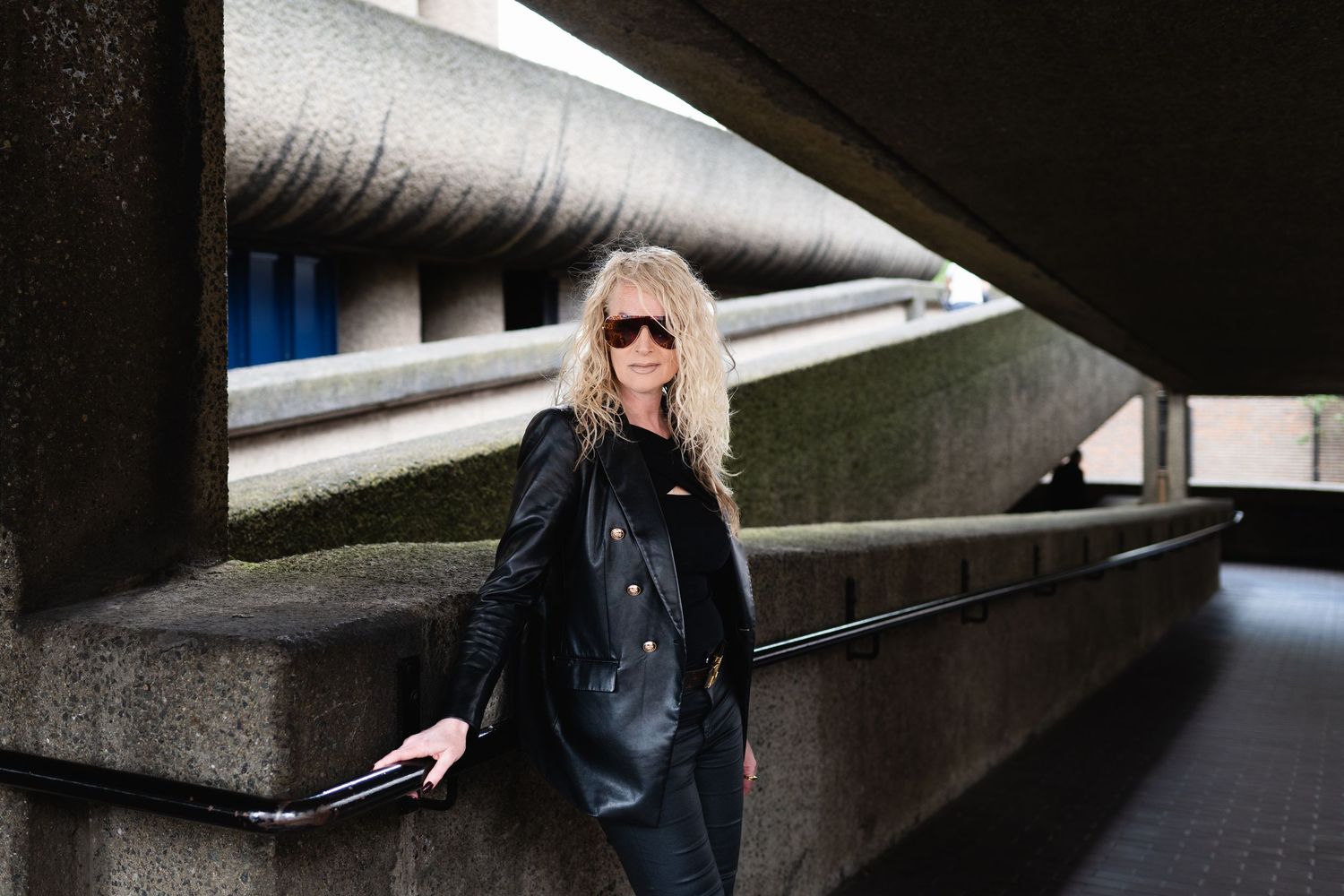 Stylish blonde woman in black leather jacket and sunglasses leaning against concrete wall outdoors.