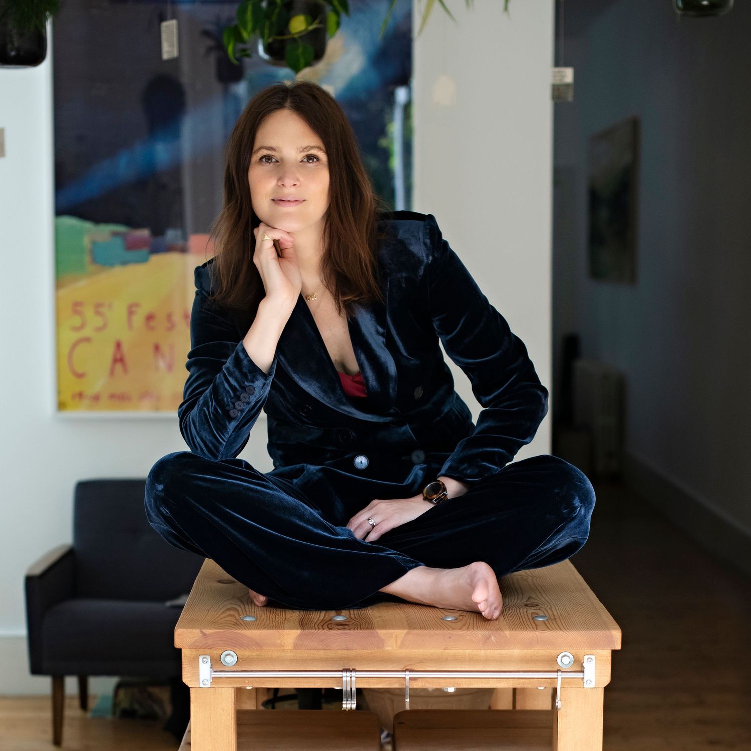 Brunette woman in dark velvet suit sitting cross-legged on a wooden table in a modern space.