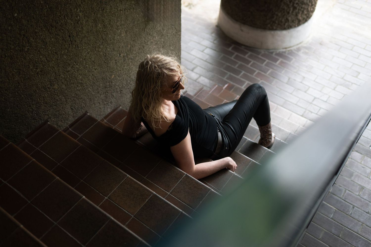Blonde woman in all-black outfit lying on brick steps in an urban architectural setting.