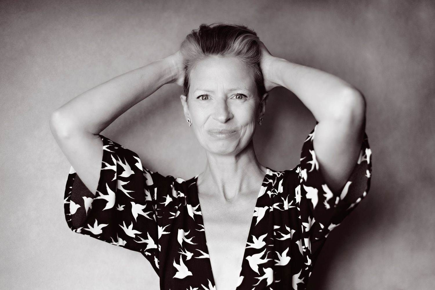Black and white portrait of woman in bird-print wrap dress with hands in hair.