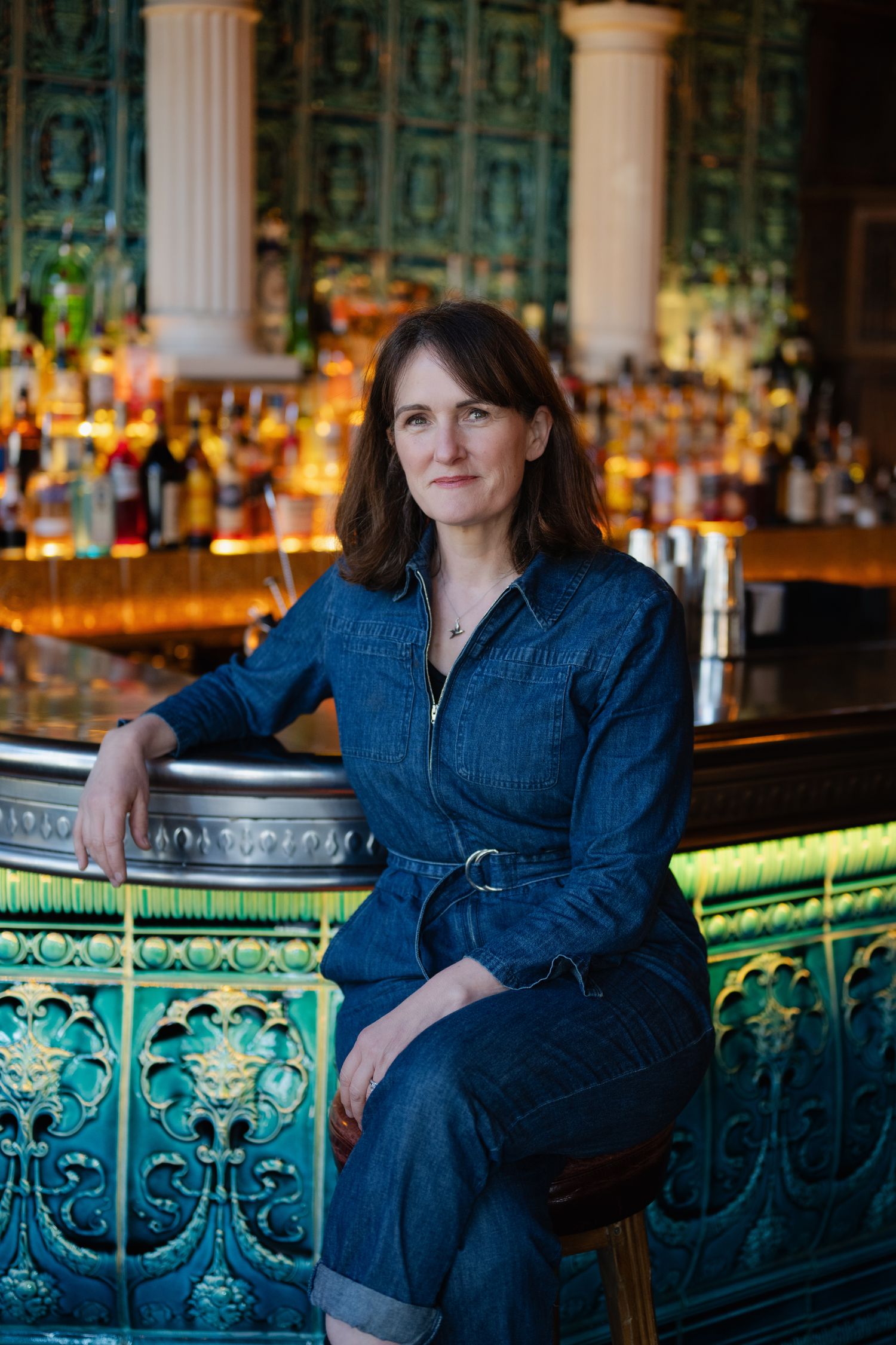 Woman in denim jumpsuit seated at ornate teal bar counter in warmly lit venue.