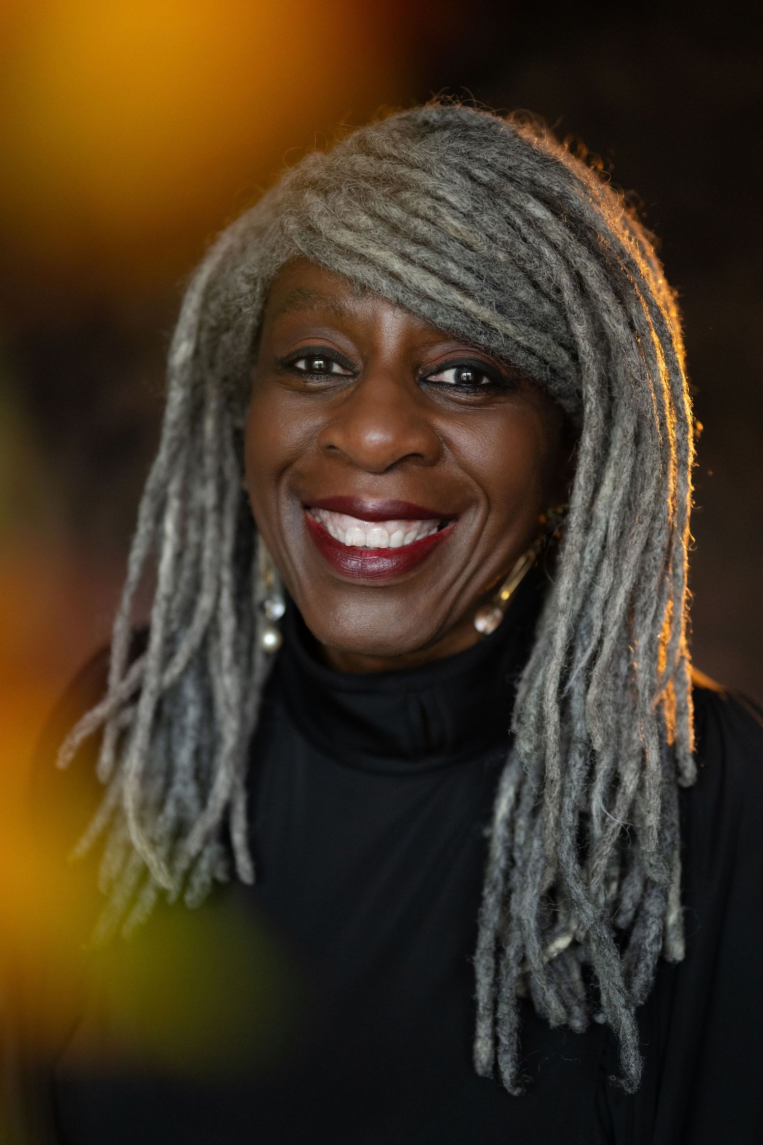 A smiling Black woman with long gray locs wearing a black top poses against a warm bokeh background.