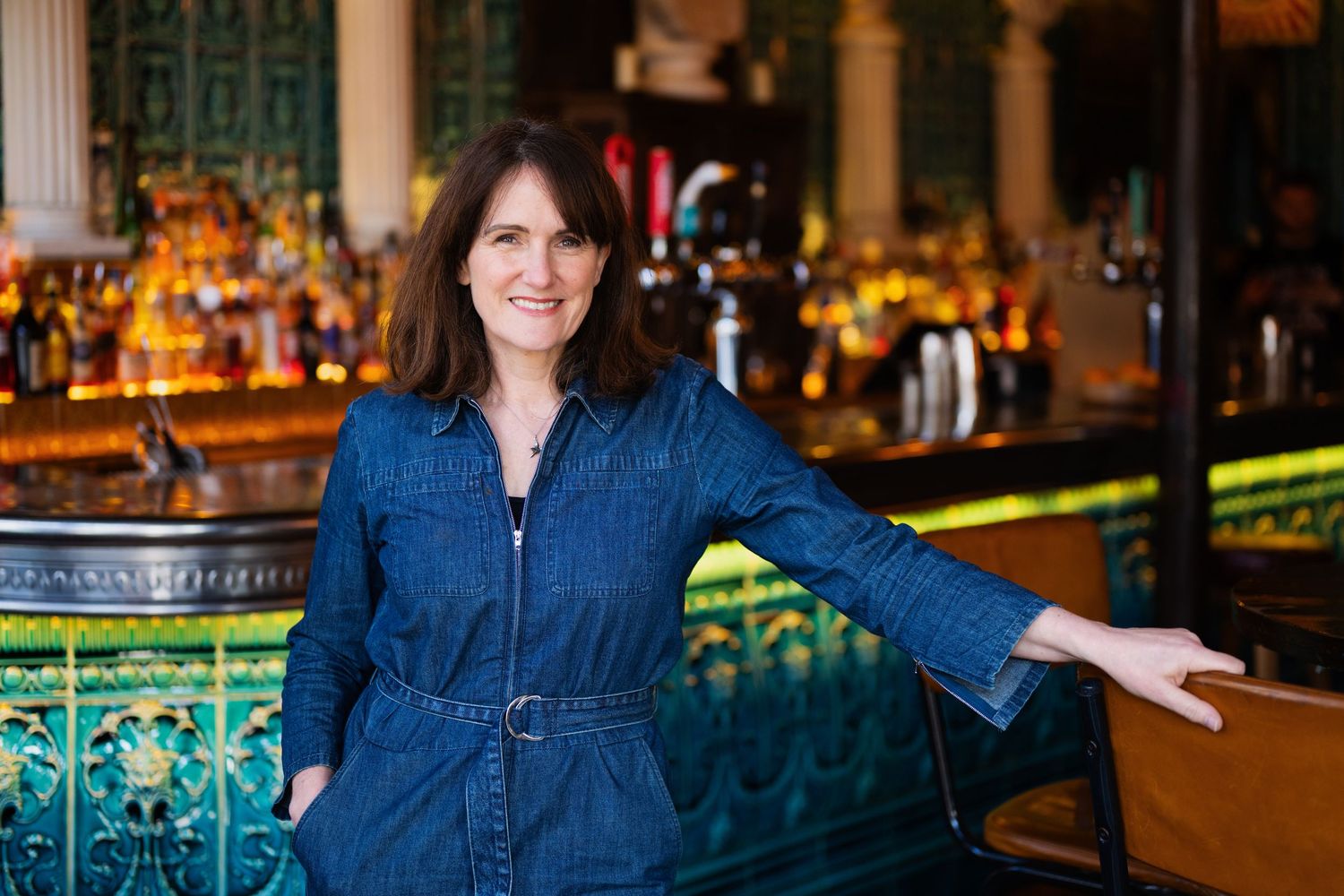 Woman in belted denim jumpsuit standing at ornate green tiled bar with bottle display.