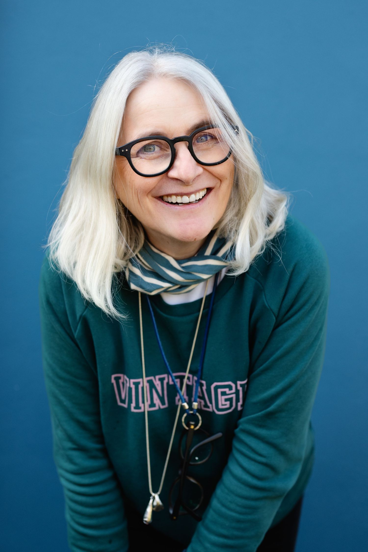 Smiling woman with white hair and glasses wearing green vintage sweatshirt on blue background.