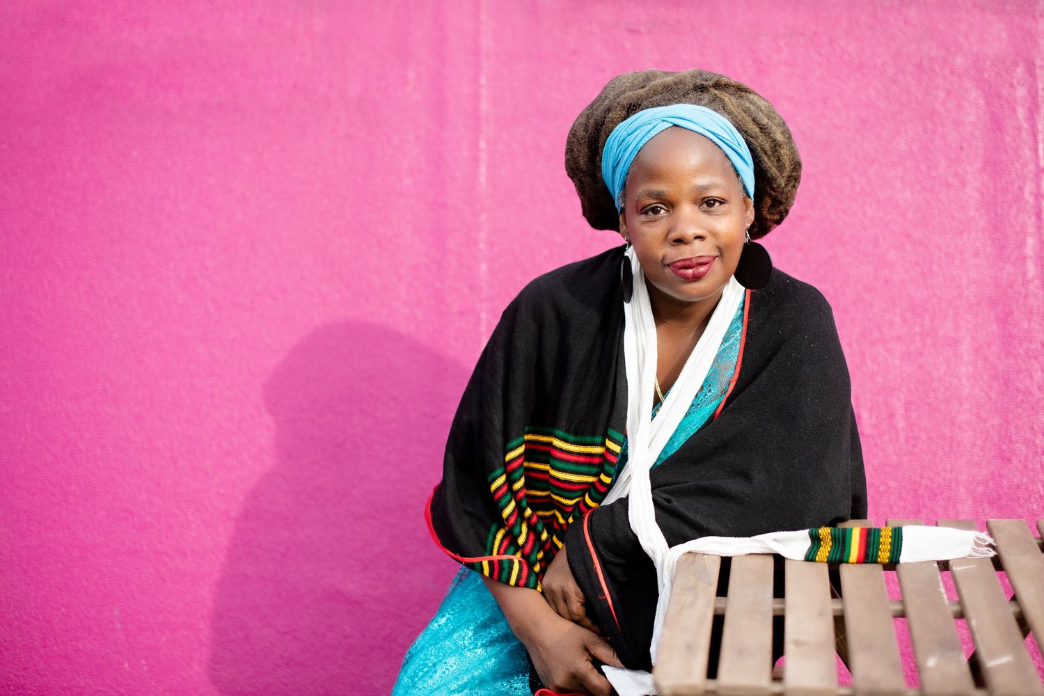 Woman in colorful African kente wrap and blue headband sits on a wooden bench against a bright pink wall.