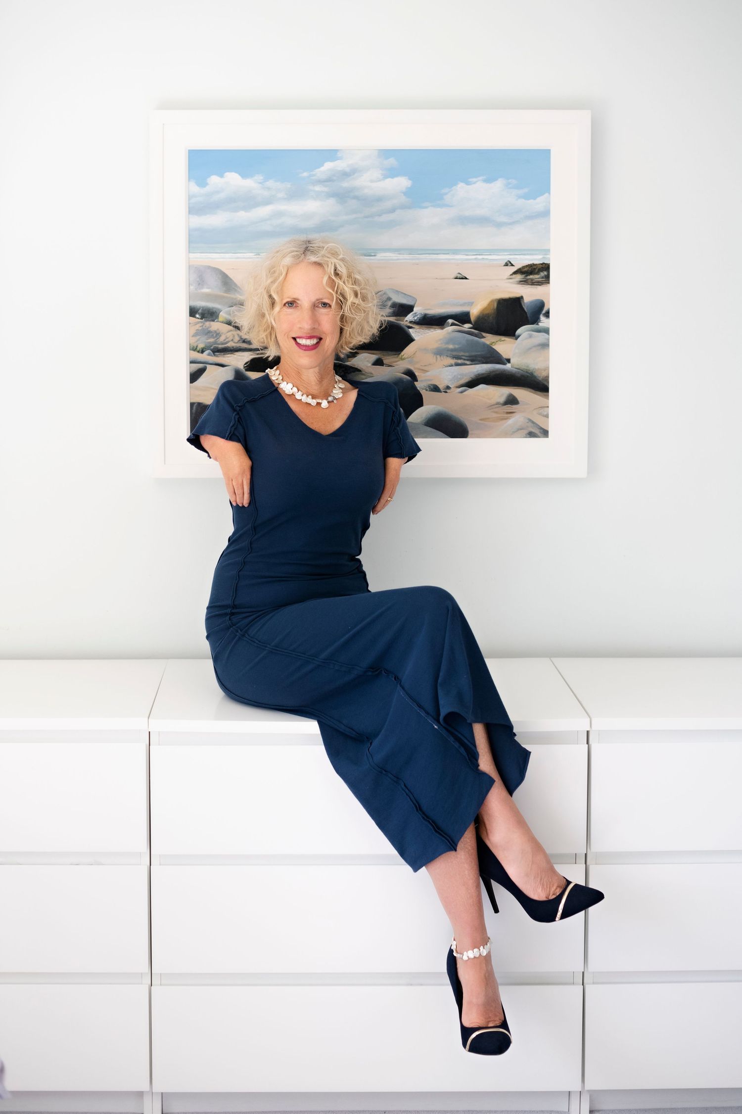 A smiling blonde woman in a navy outfit and pearl necklace sits on a white dresser in front of a coastal landscape painting.