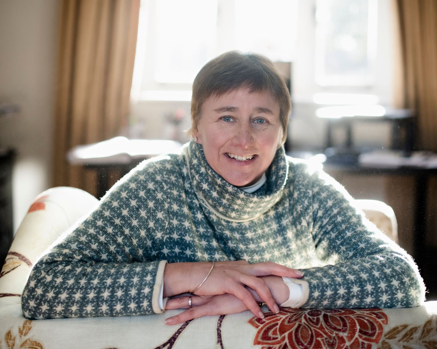 Smiling middle-aged woman in a teal patterned sweater leaning on a floral chair in a cozy room.