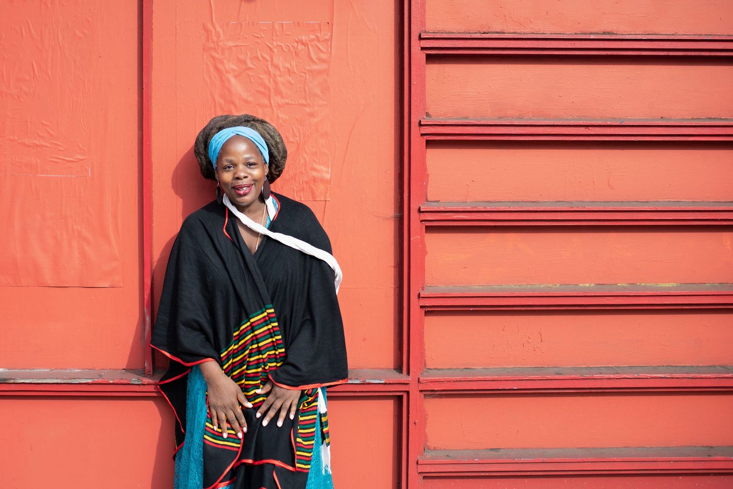 Smiling woman in traditional African black cape and kente cloth leans against a red wall in natural light.