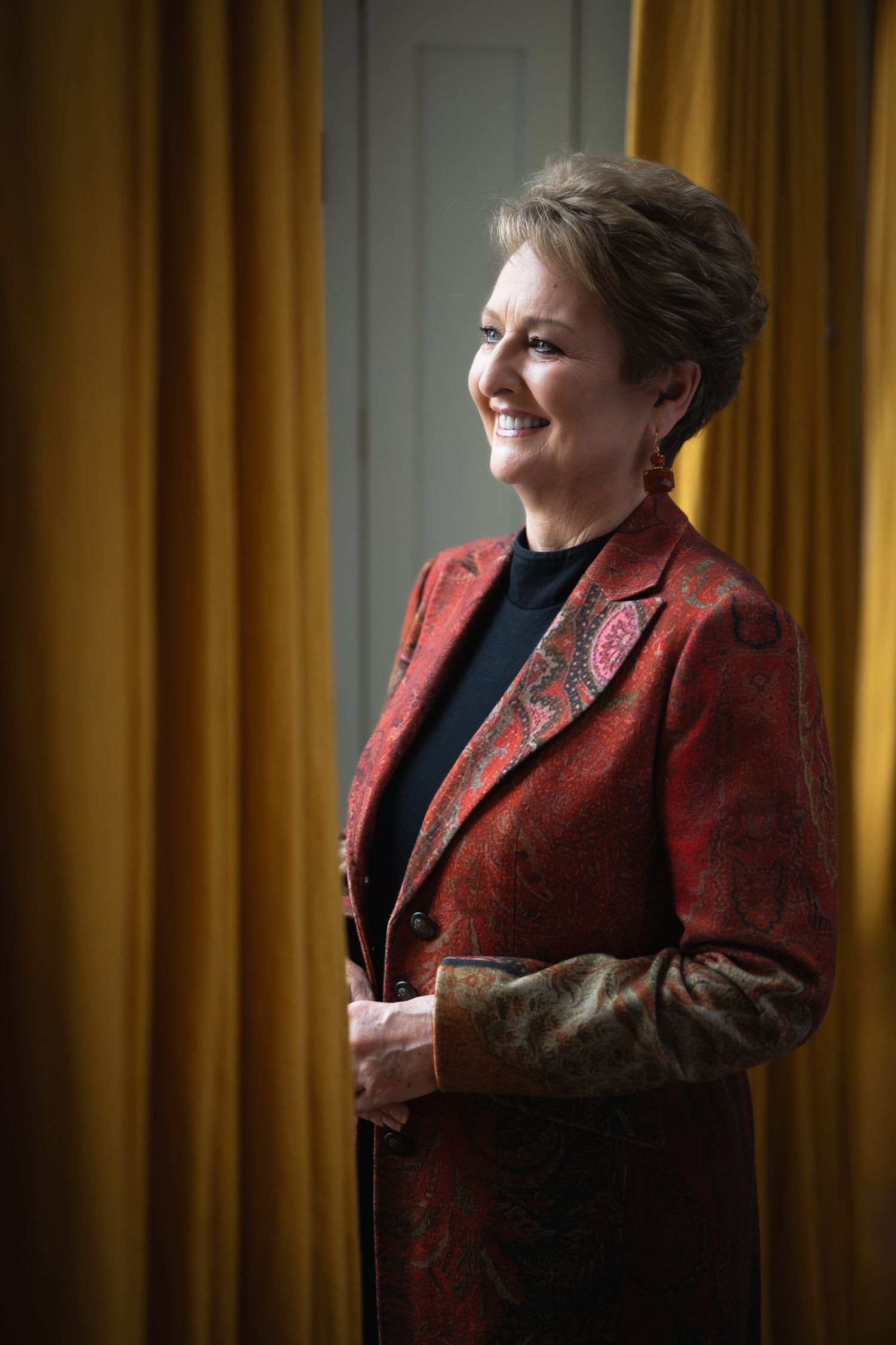 Smiling woman in a red paisley blazer standing by golden curtains in elegant interior.