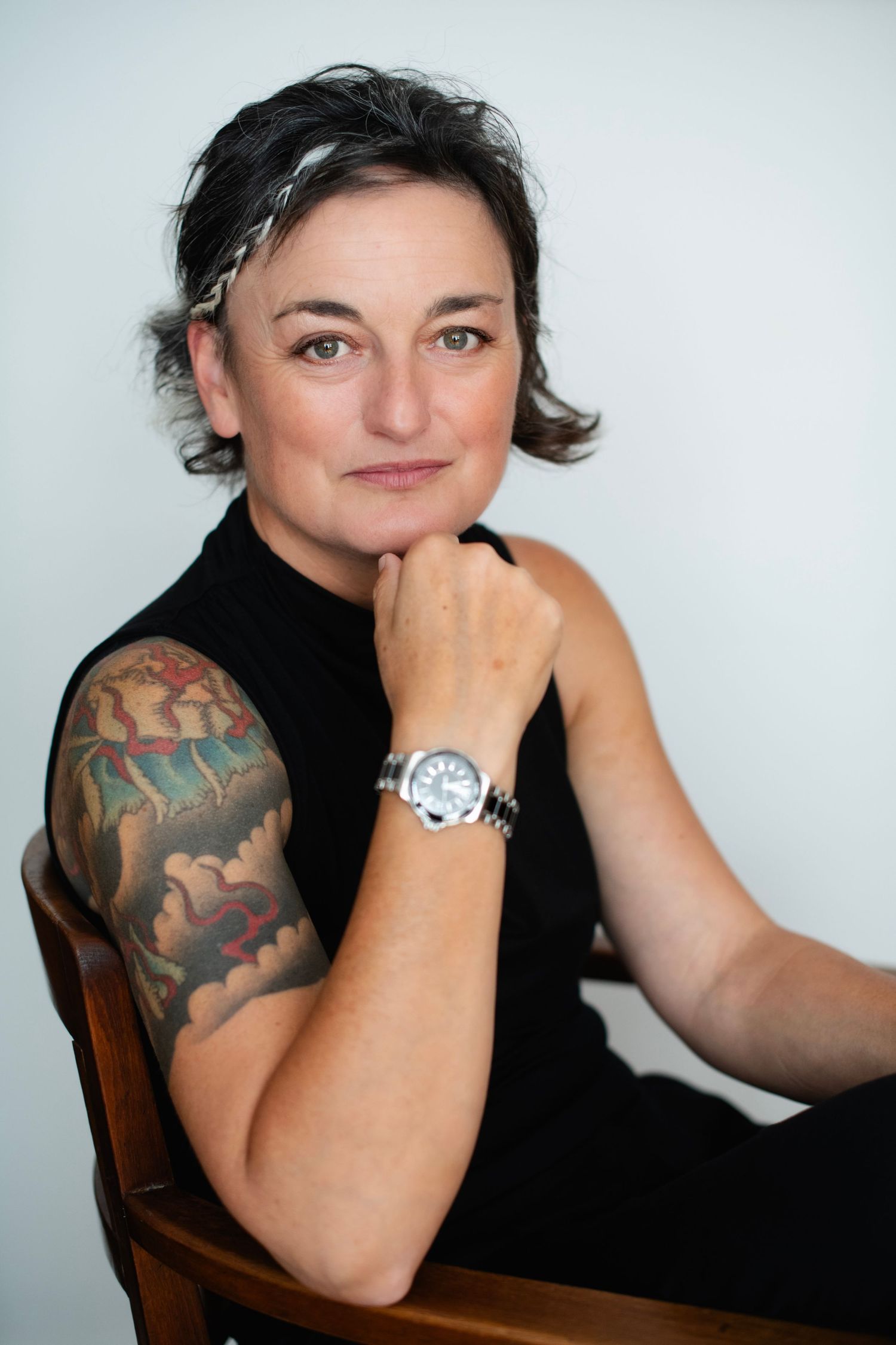 Tattooed woman in black sleeveless top seated on wooden chair, resting chin on hand, wearing silver watch.