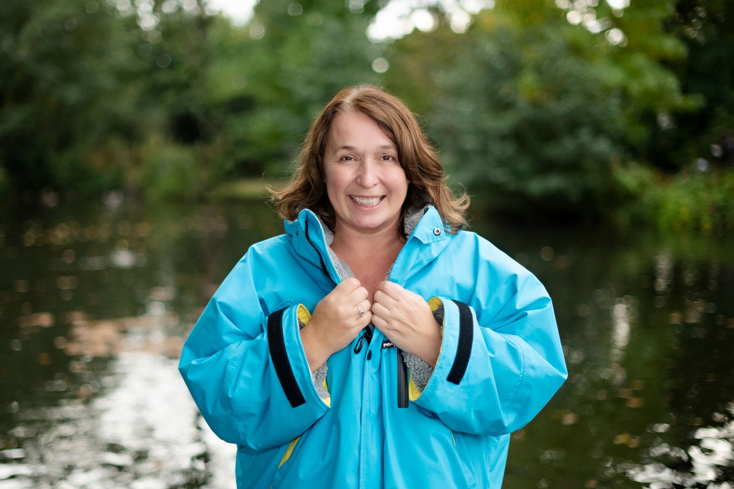 Woman smiling in a turquoise rain jacket standing by a river with lush green trees in the background.