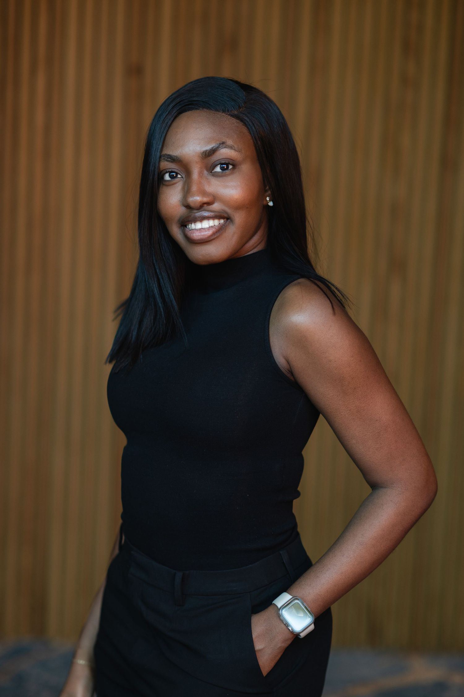 Smiling Black woman in black sleeveless turtleneck and pants, posing confidently with hand in pocket against wooden background.