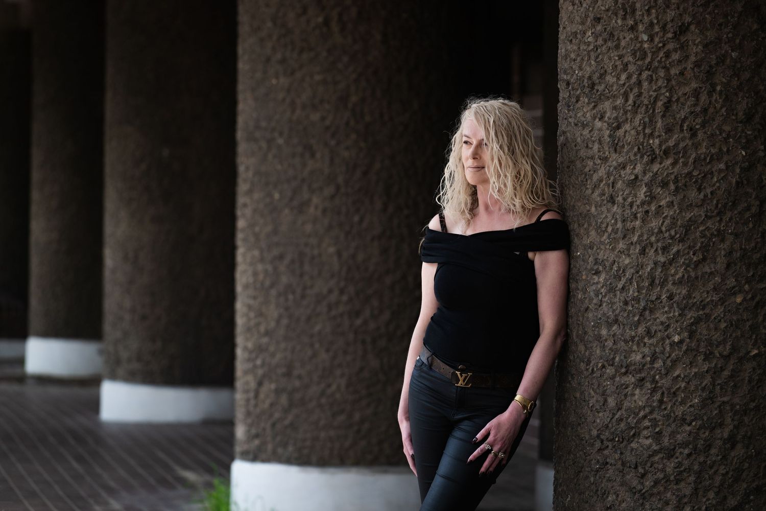 Blonde woman in black off-shoulder top leans against textured columns in an outdoor architectural setting.