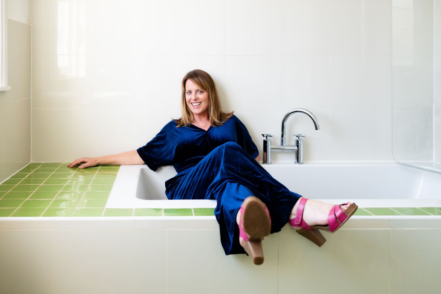 A cheerful woman in a blue velvet outfit sits on the edge of a white bathtub with green tile accents.