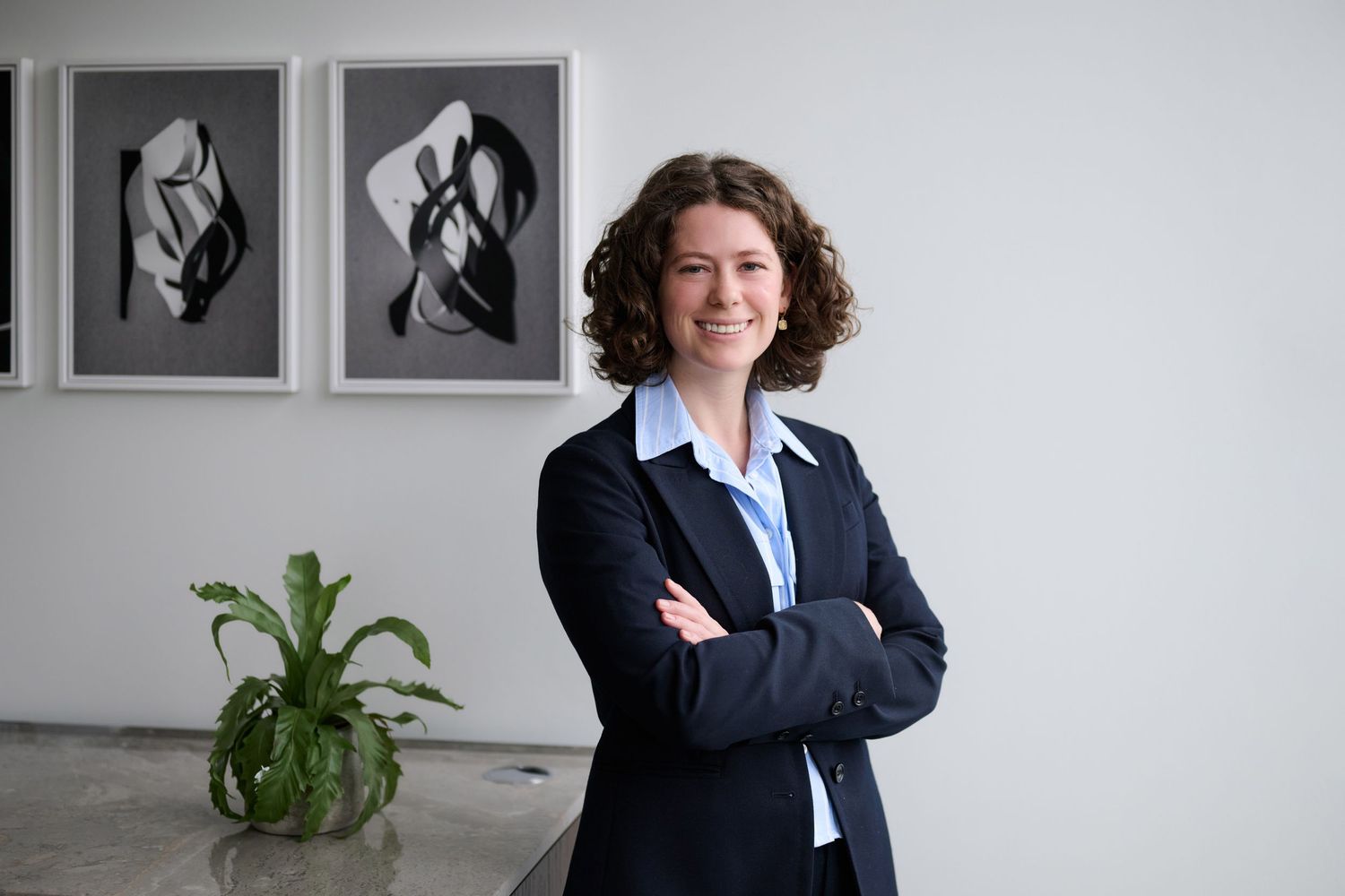 Smiling woman in navy blazer standing with arms crossed beside plant and abstract black and white artwork.