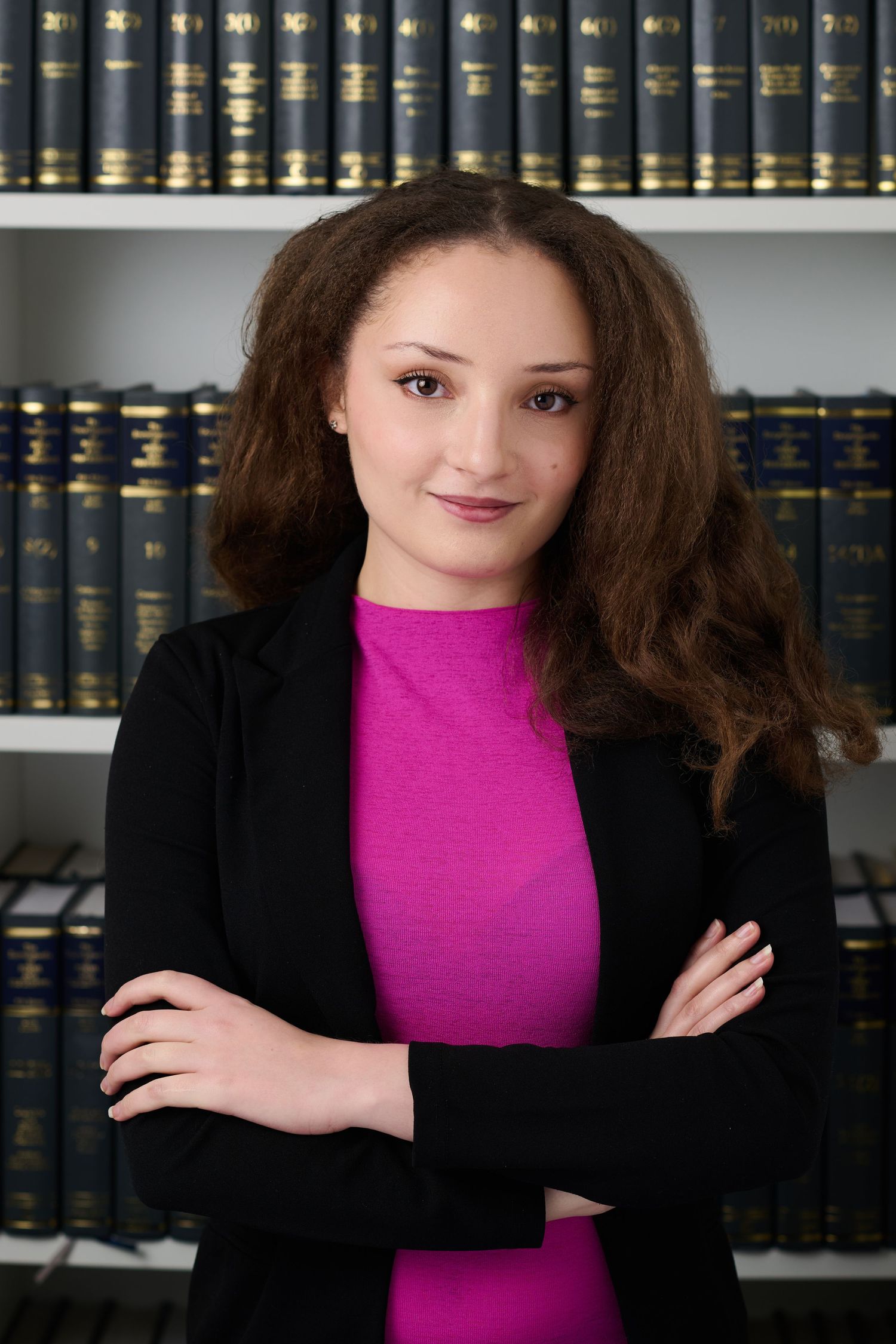 Young professional woman with curly brown hair stands arms crossed before law book shelves.