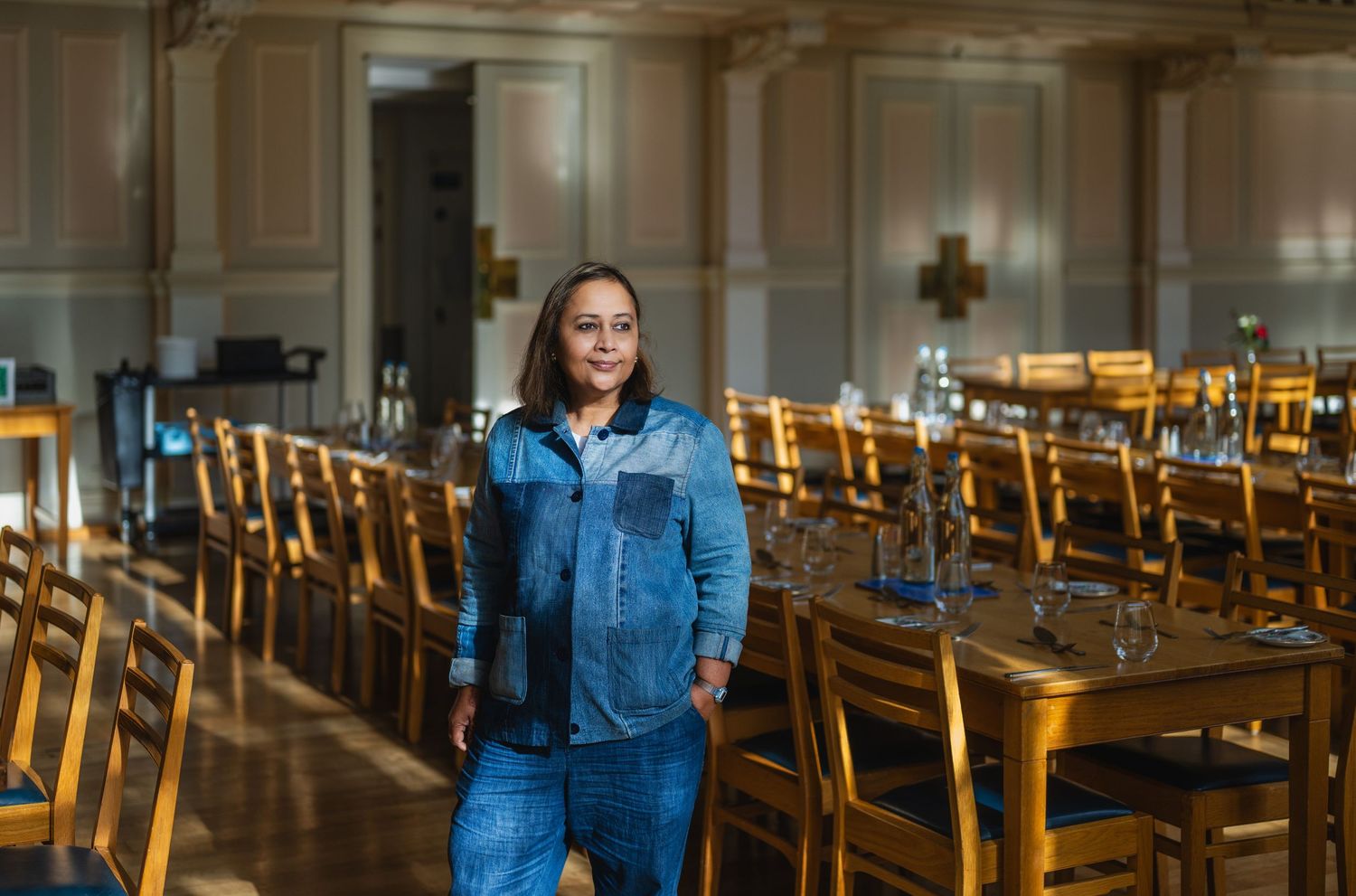 A woman in a denim jacket stands confidently in an elegant dining hall with rows of wooden chairs.