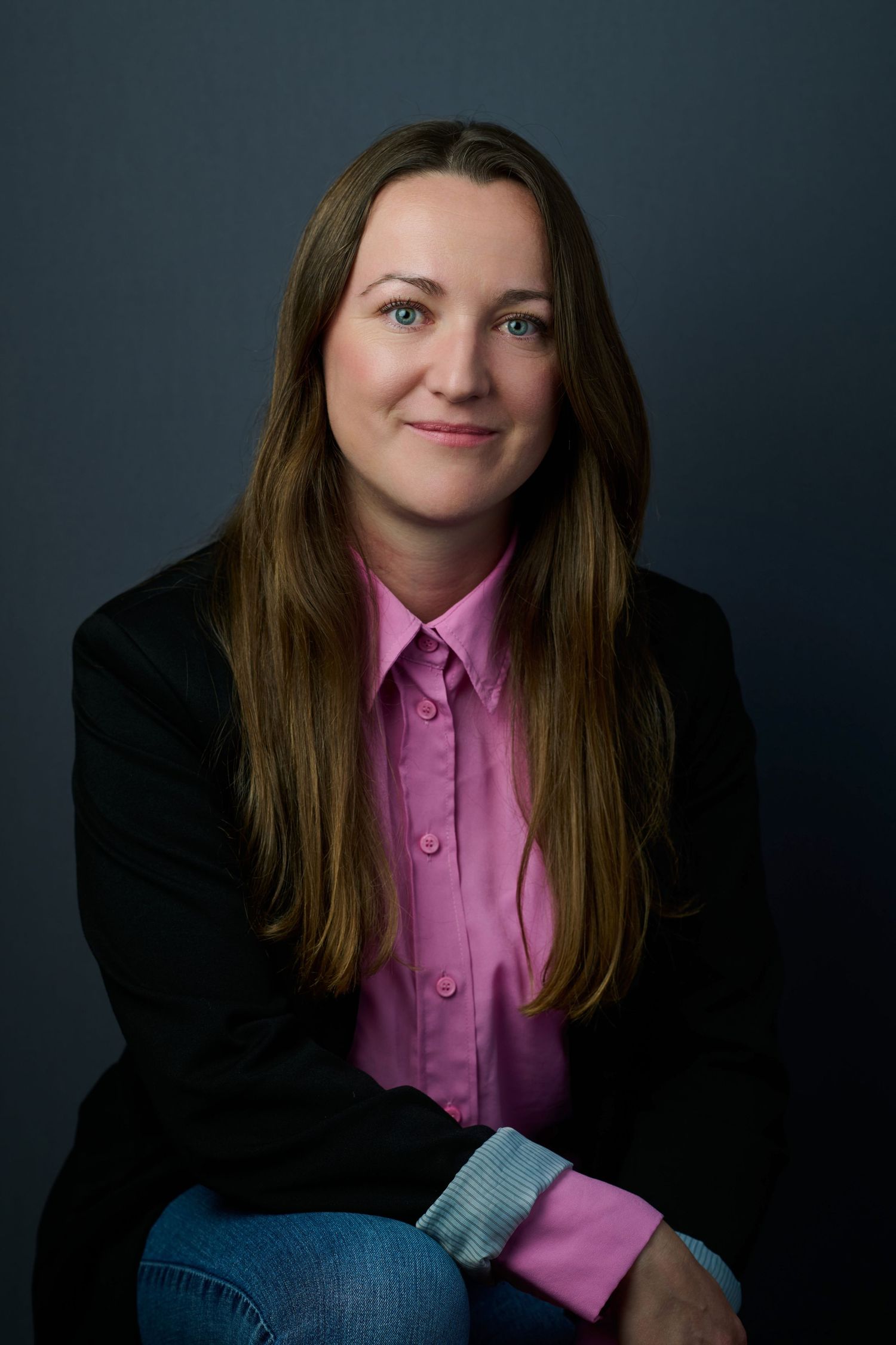 Woman with long brown hair in pink shirt and black blazer sitting against a blue-gray background.