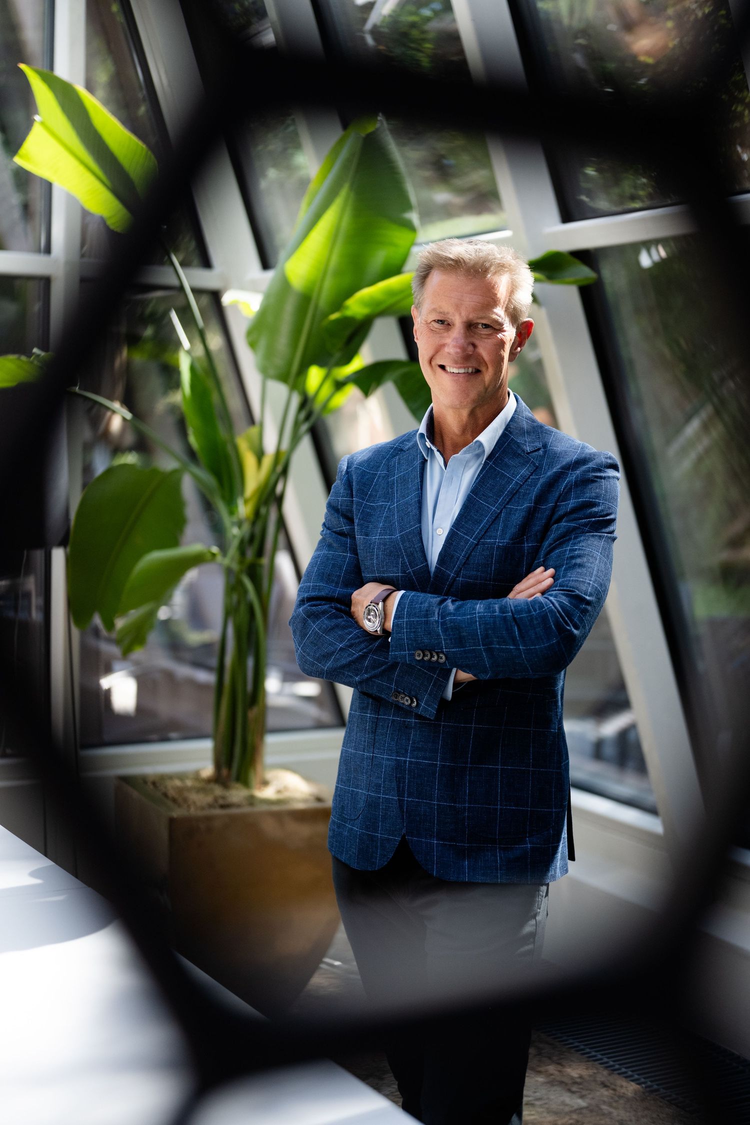 Smiling man in blue plaid blazer with arms crossed, photographed through geometric frame with tropical plants.