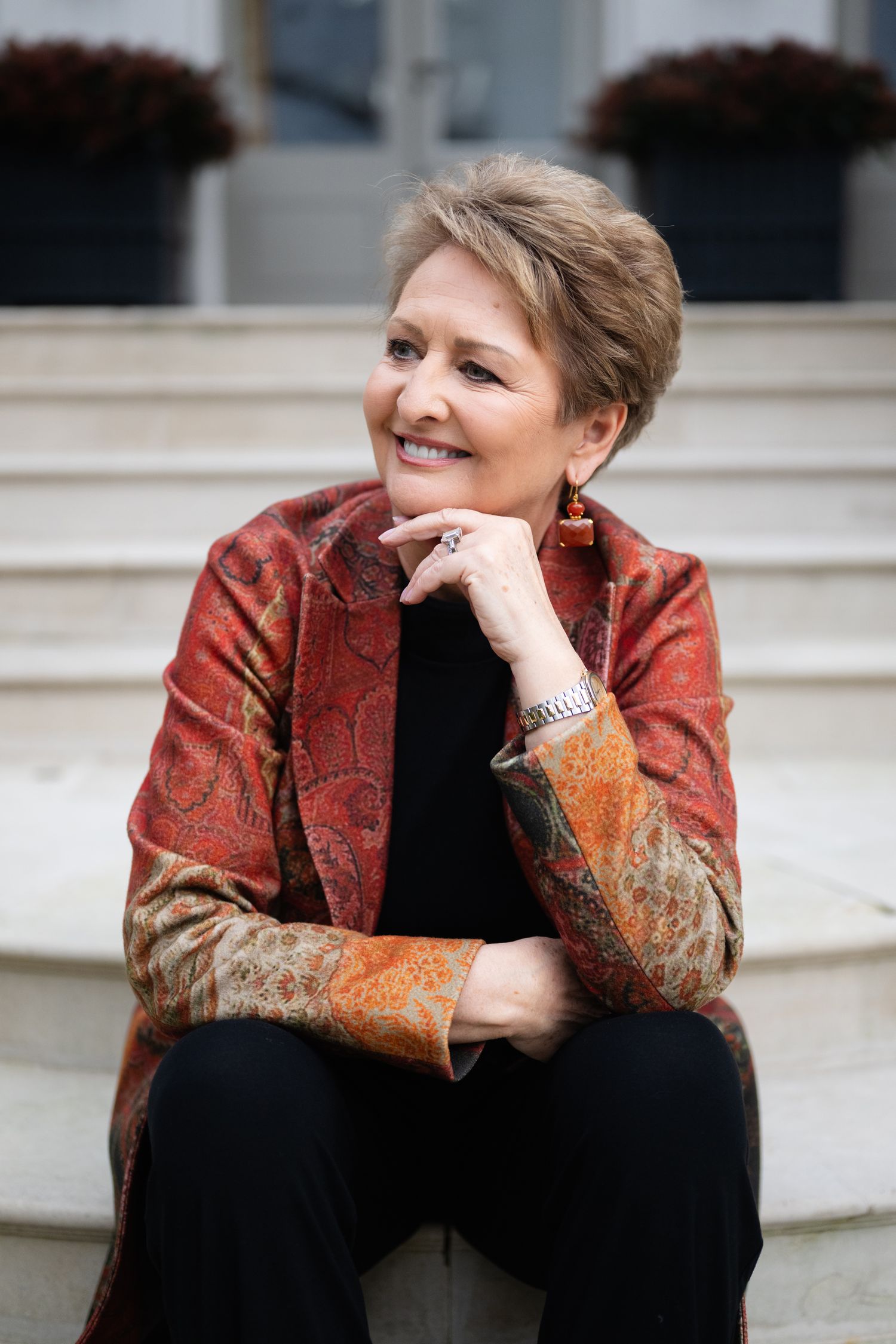 Smiling woman in red paisley jacket sitting on stone steps with hand on chin.