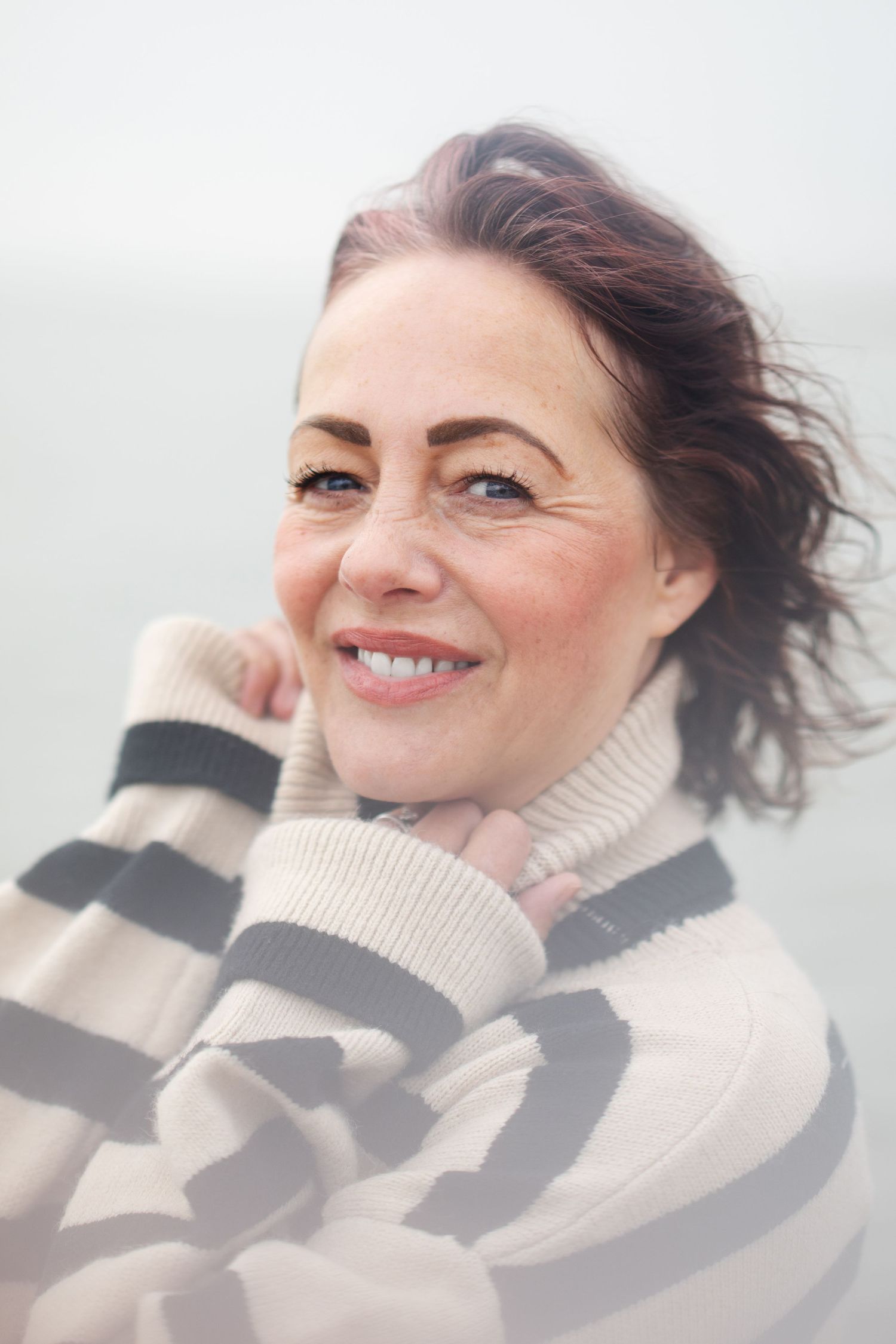 A smiling middle-aged woman in a striped turtleneck sweater poses on a windy beach.
