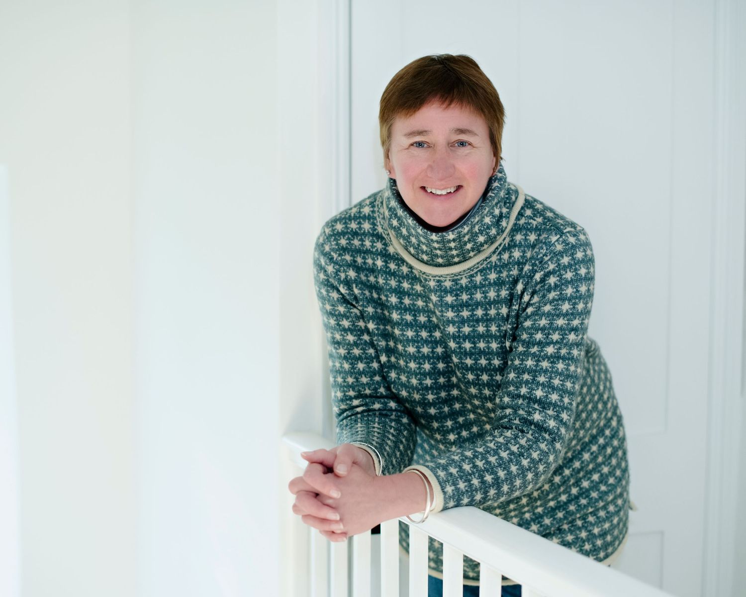 Cheerful woman in a green patterned turtleneck sweater leaning on a white railing indoors.
