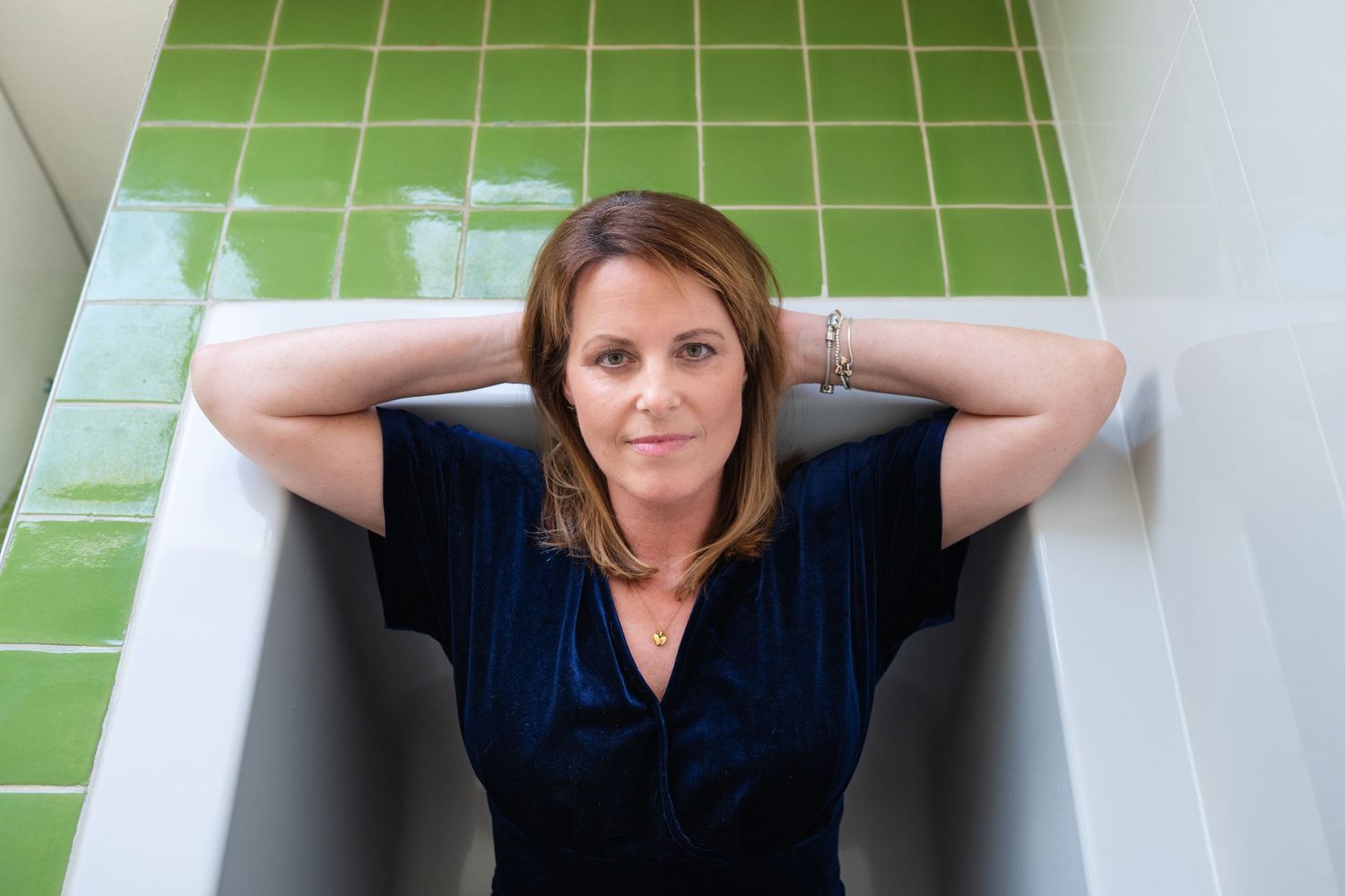 Woman in blue velvet top relaxing in bathtub with green tiled walls, hands behind head.
