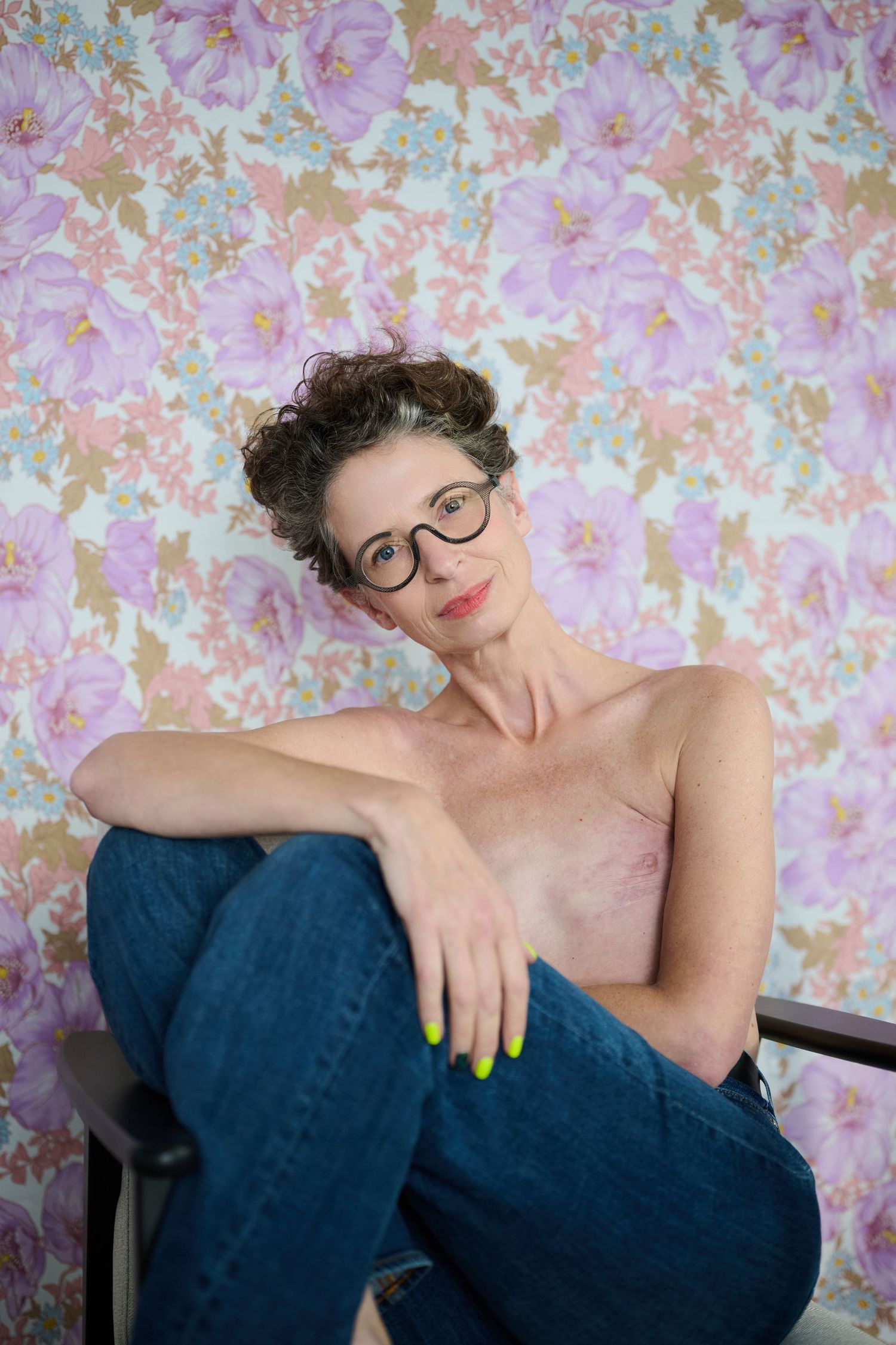 Woman with curly hair and round glasses sitting in jeans against floral wallpaper.