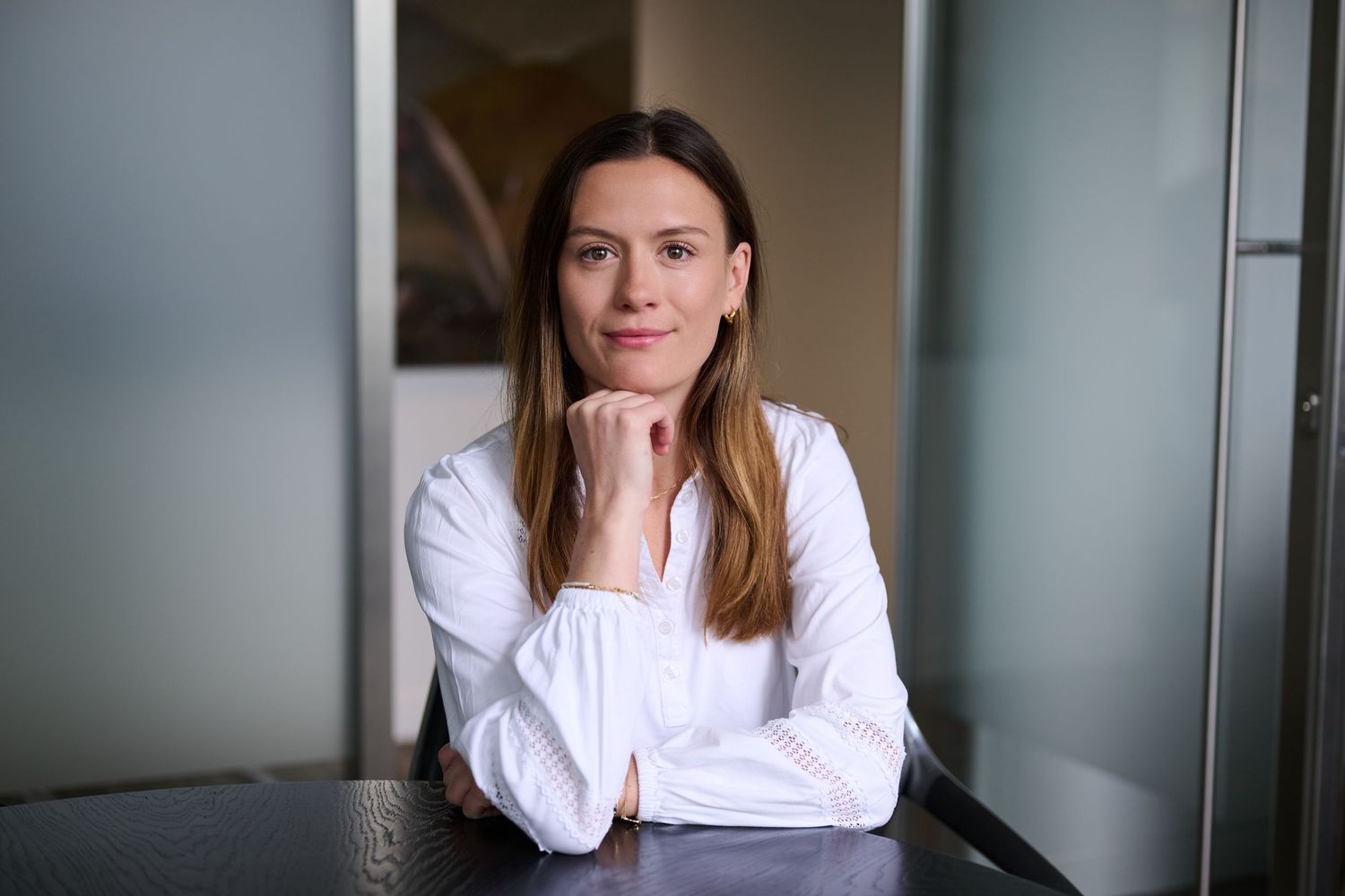 Confident woman in white blouse seated at dark table with chin resting on hands in contemporary office.