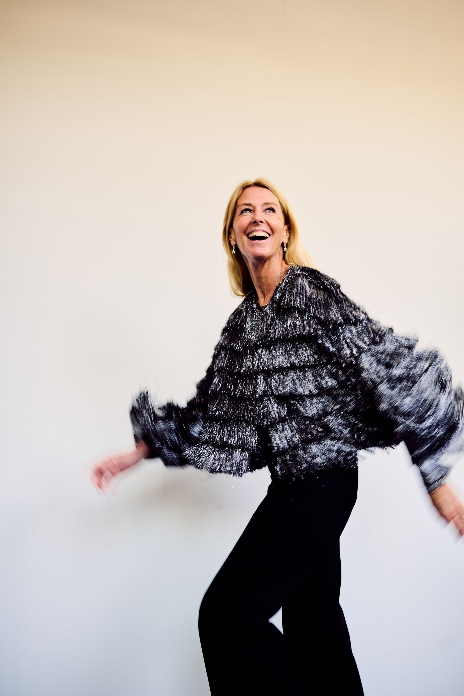 Joyful blonde woman in textured black and silver jacket laughs and dances against a neutral background.