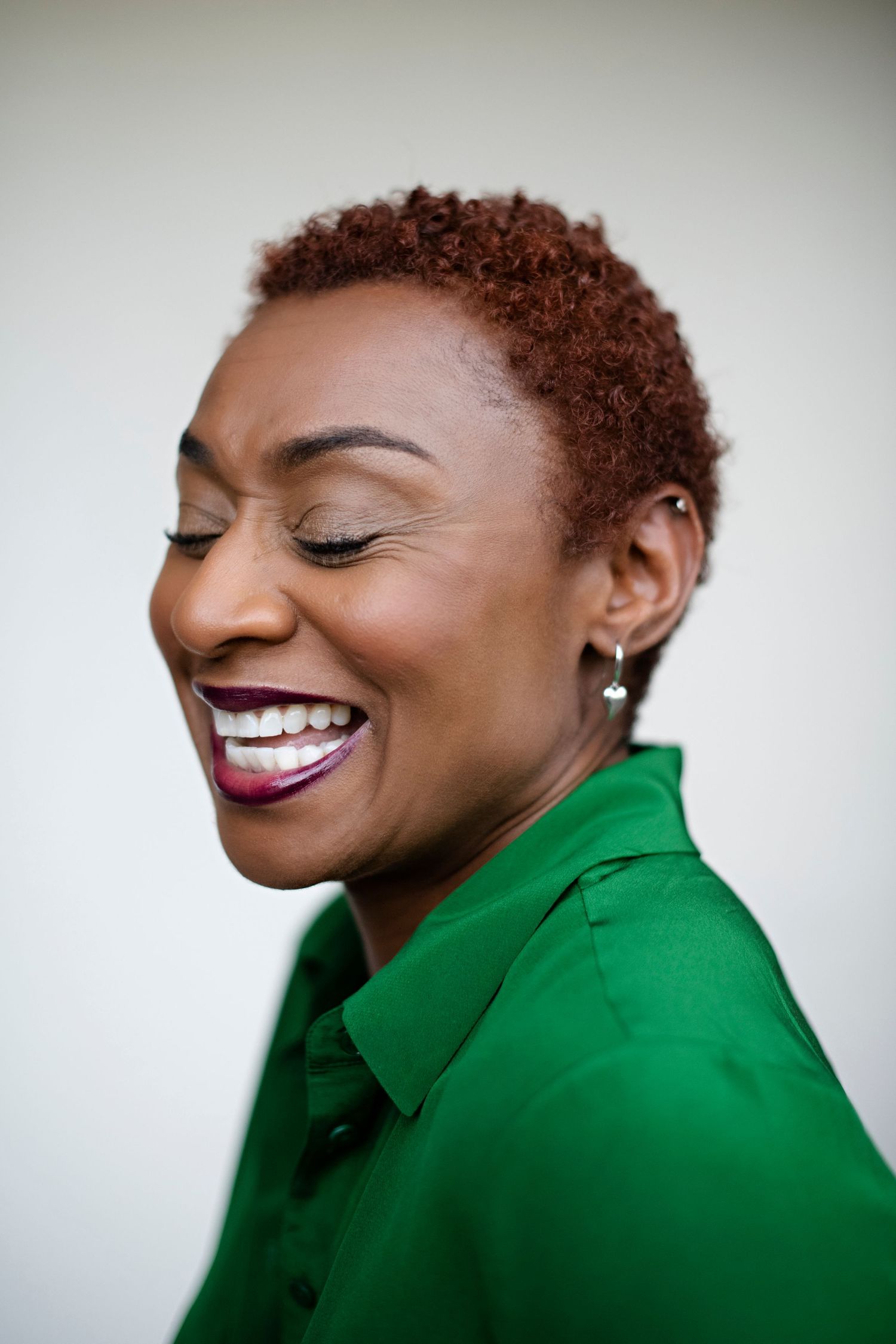 Black woman with short auburn hair laughing with eyes closed wearing a bright green shirt and silver drop earrings.