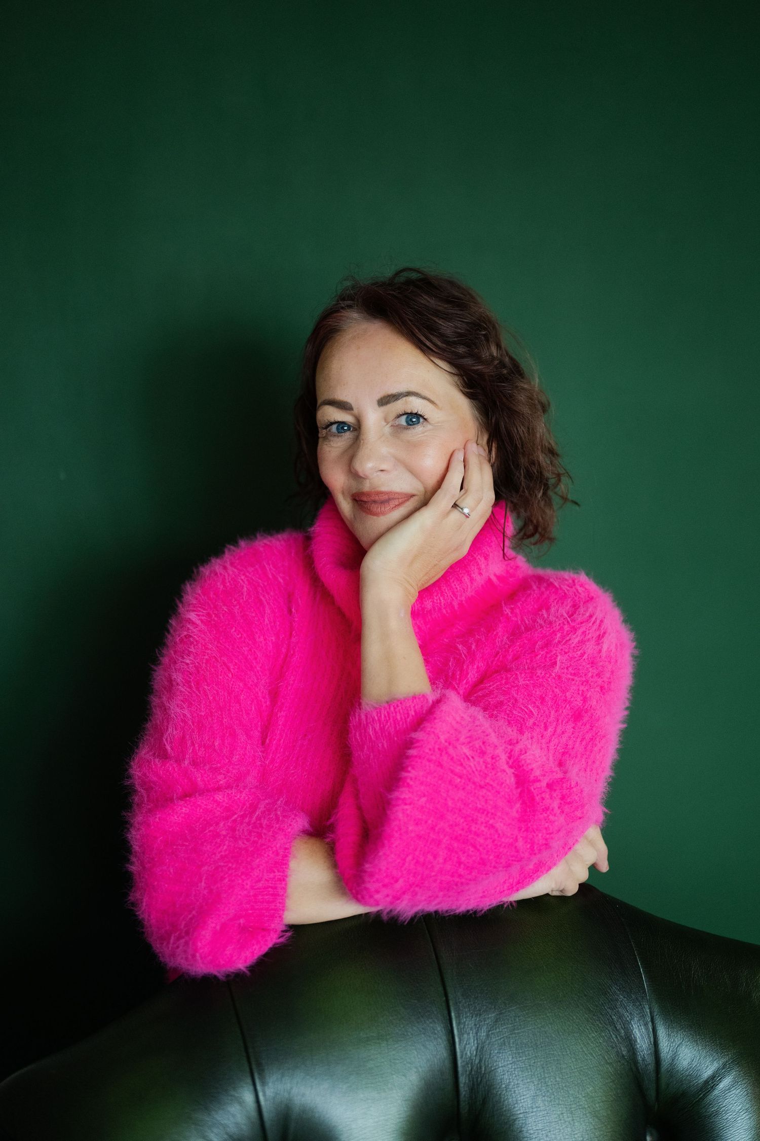 Woman in bright pink fluffy sweater sitting on leather sofa against dark green wall, smiling.