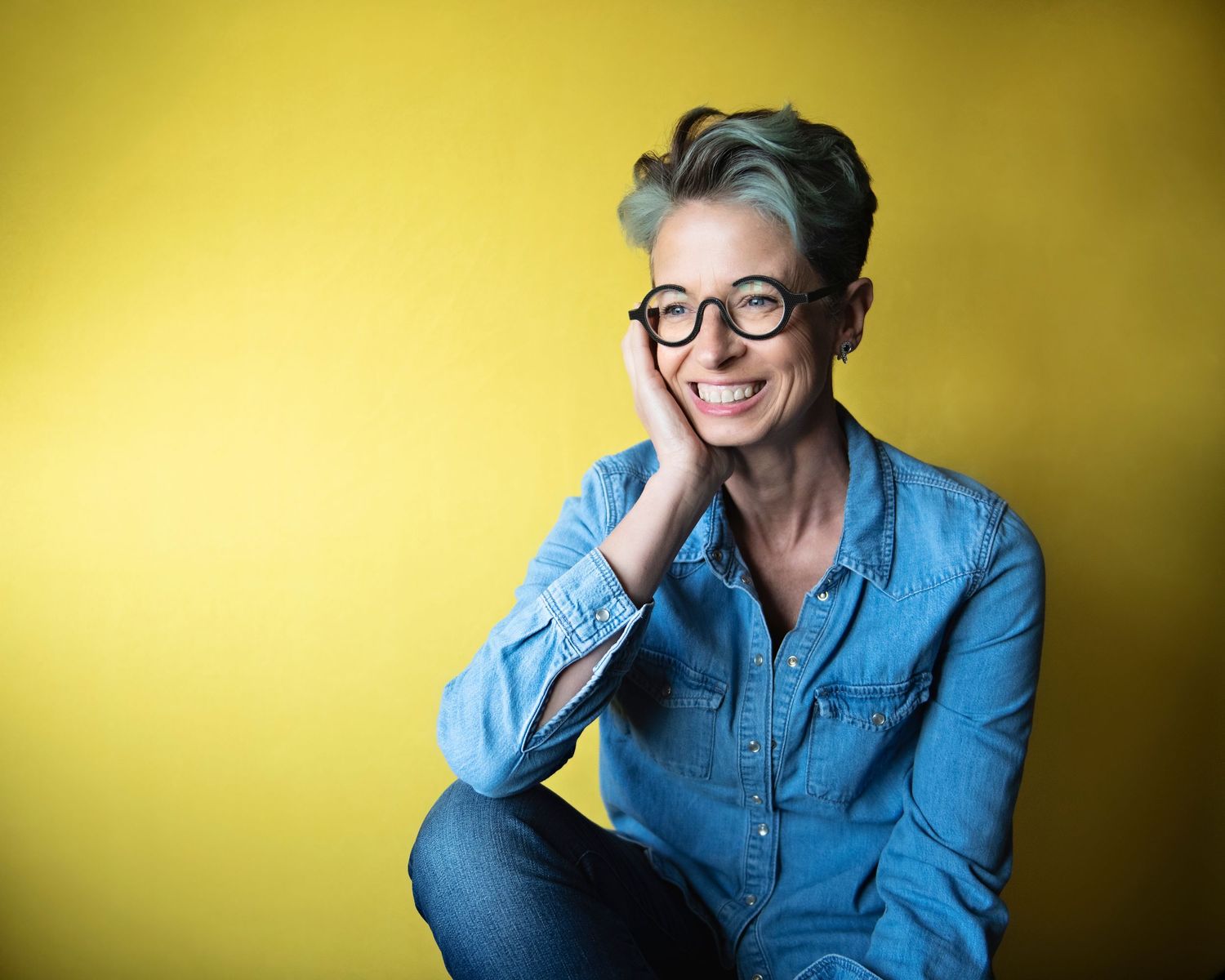 Smiling woman with short gray-green hair and round glasses in a blue denim shirt against a yellow background.