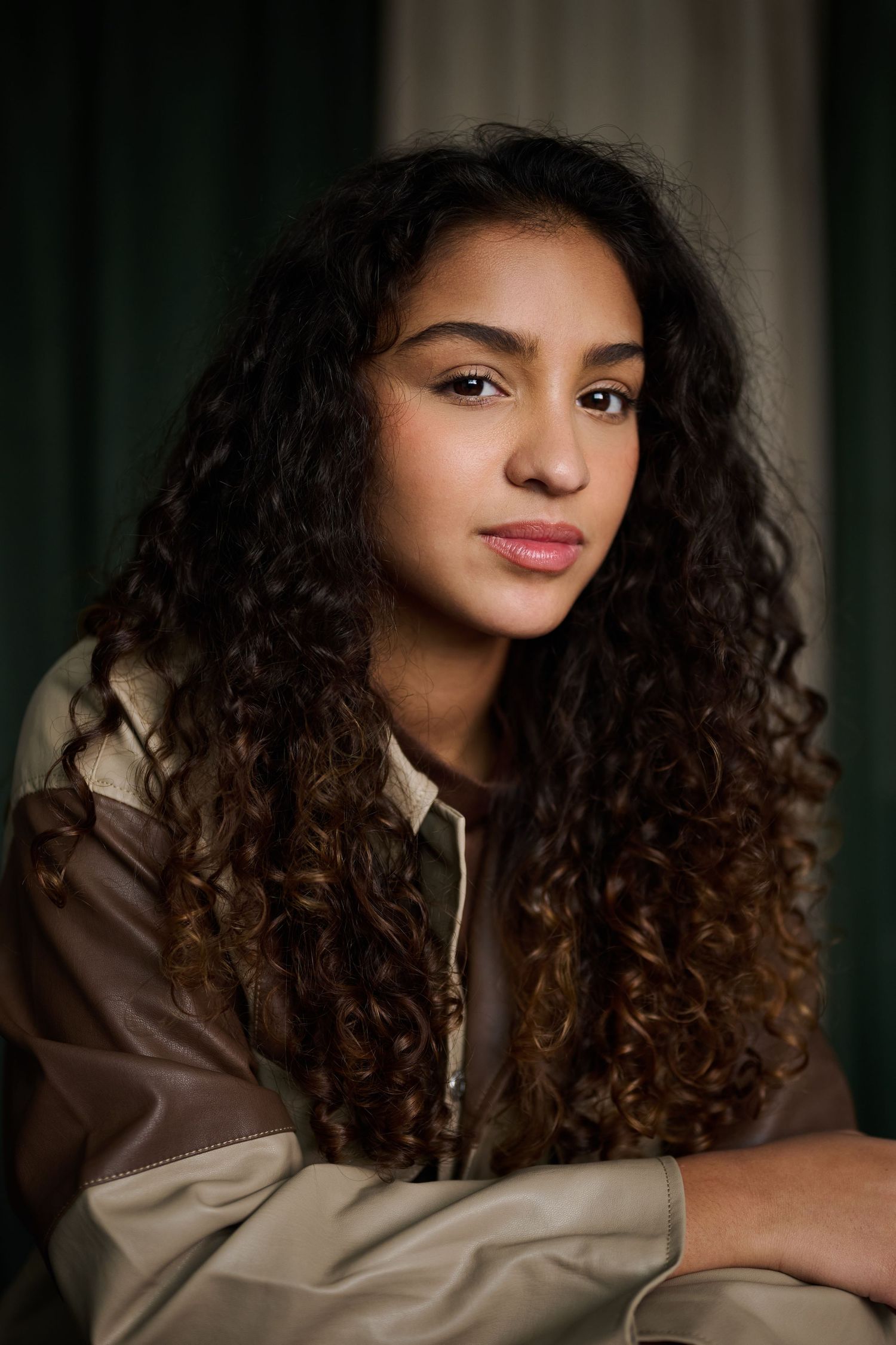 Young woman with long curly dark hair wearing a brown leather jacket, posing confidently against a green backdrop.
