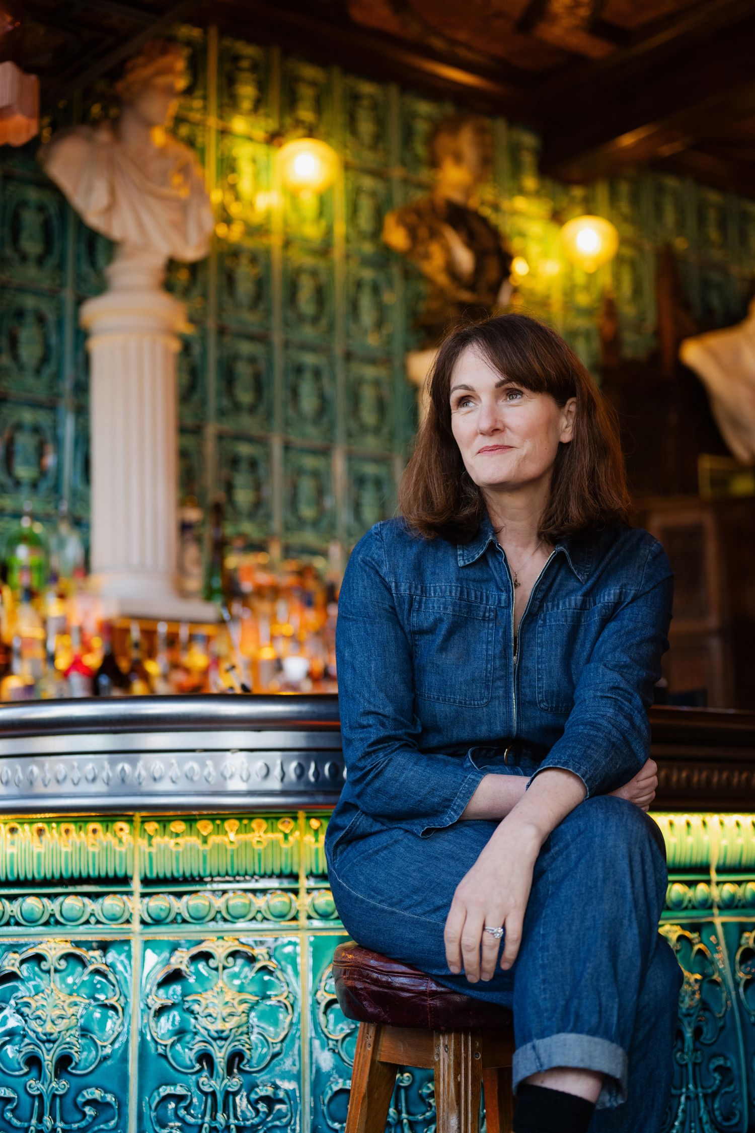 Woman in denim jumpsuit seated on ornate tiled bar stool in decorative green tiled pub interior.