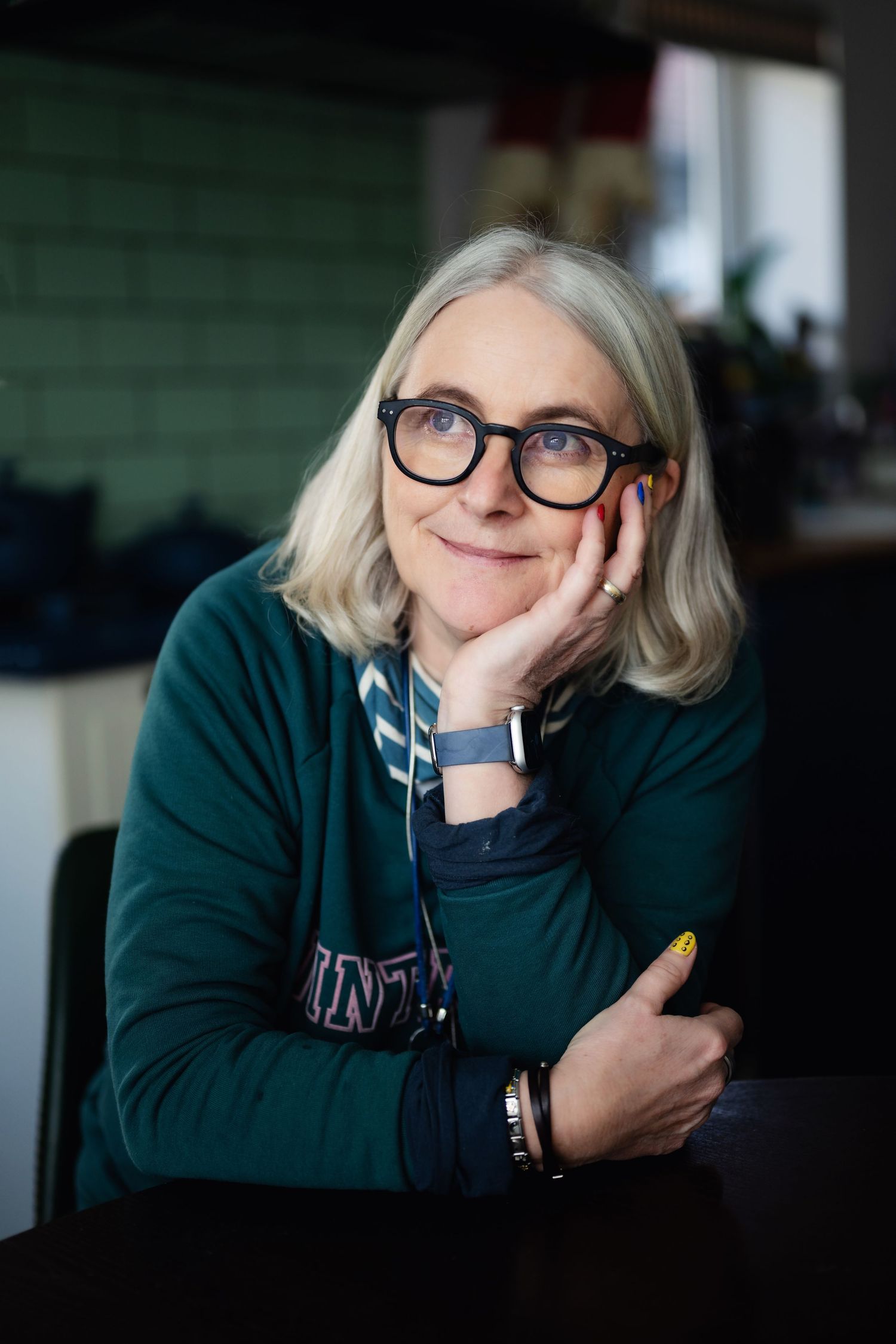 Smiling woman with gray bob haircut and black glasses rests chin on hand at a table.