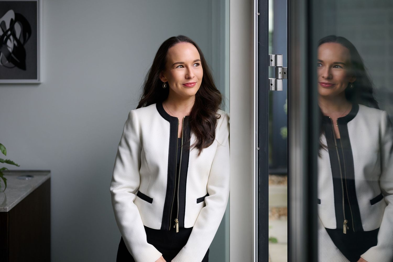 Elegant woman in white blazer gazing through glass door with reflection visible in modern office.