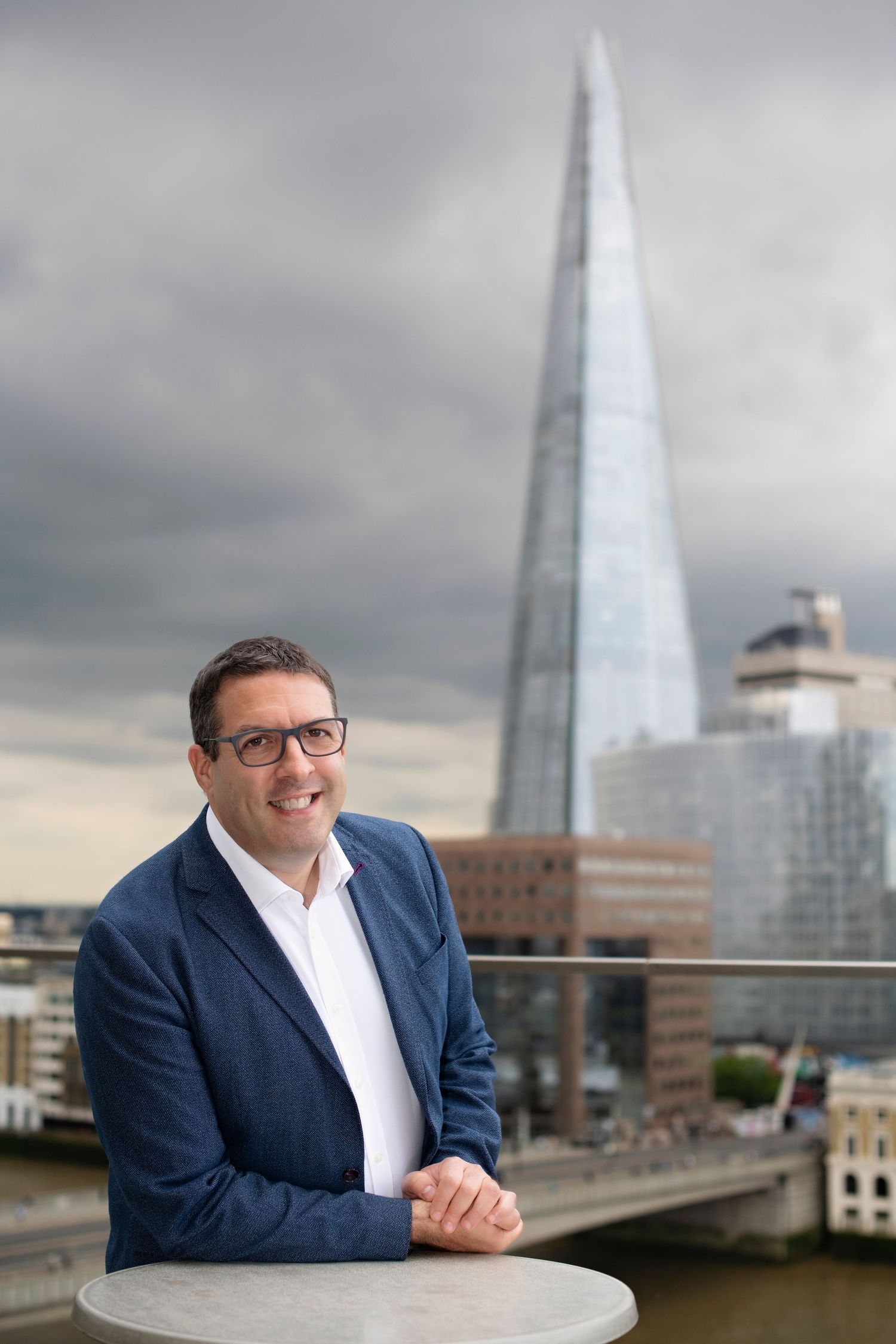 Man in blue blazer smiling on rooftop terrace with The Shard skyscraper in London background.