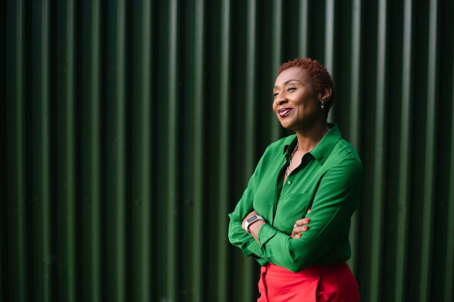 Confident woman in green jacket stands arms crossed against green corrugated wall.