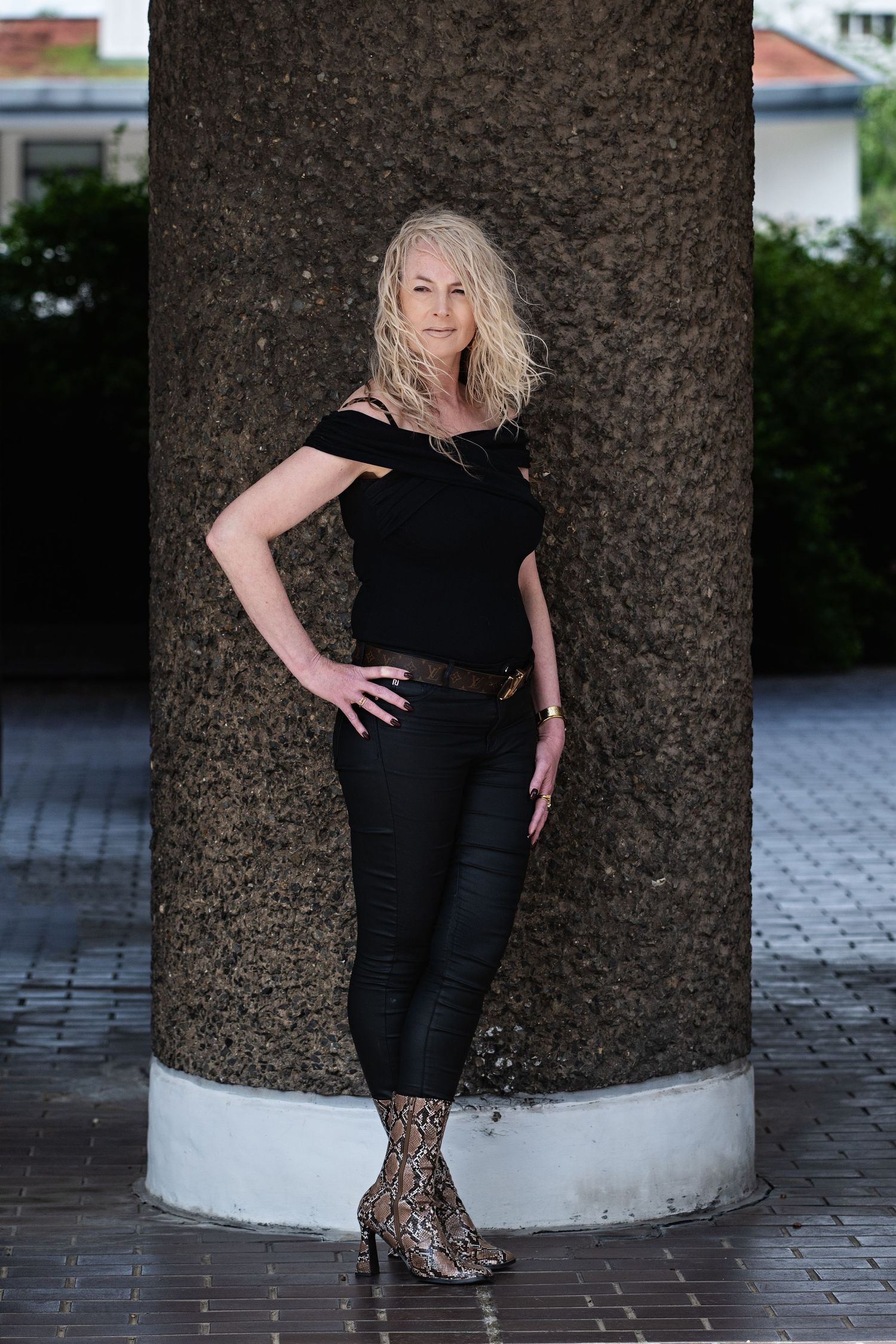 Blonde woman in black off-shoulder top and snake-print boots leaning against a large stone column.