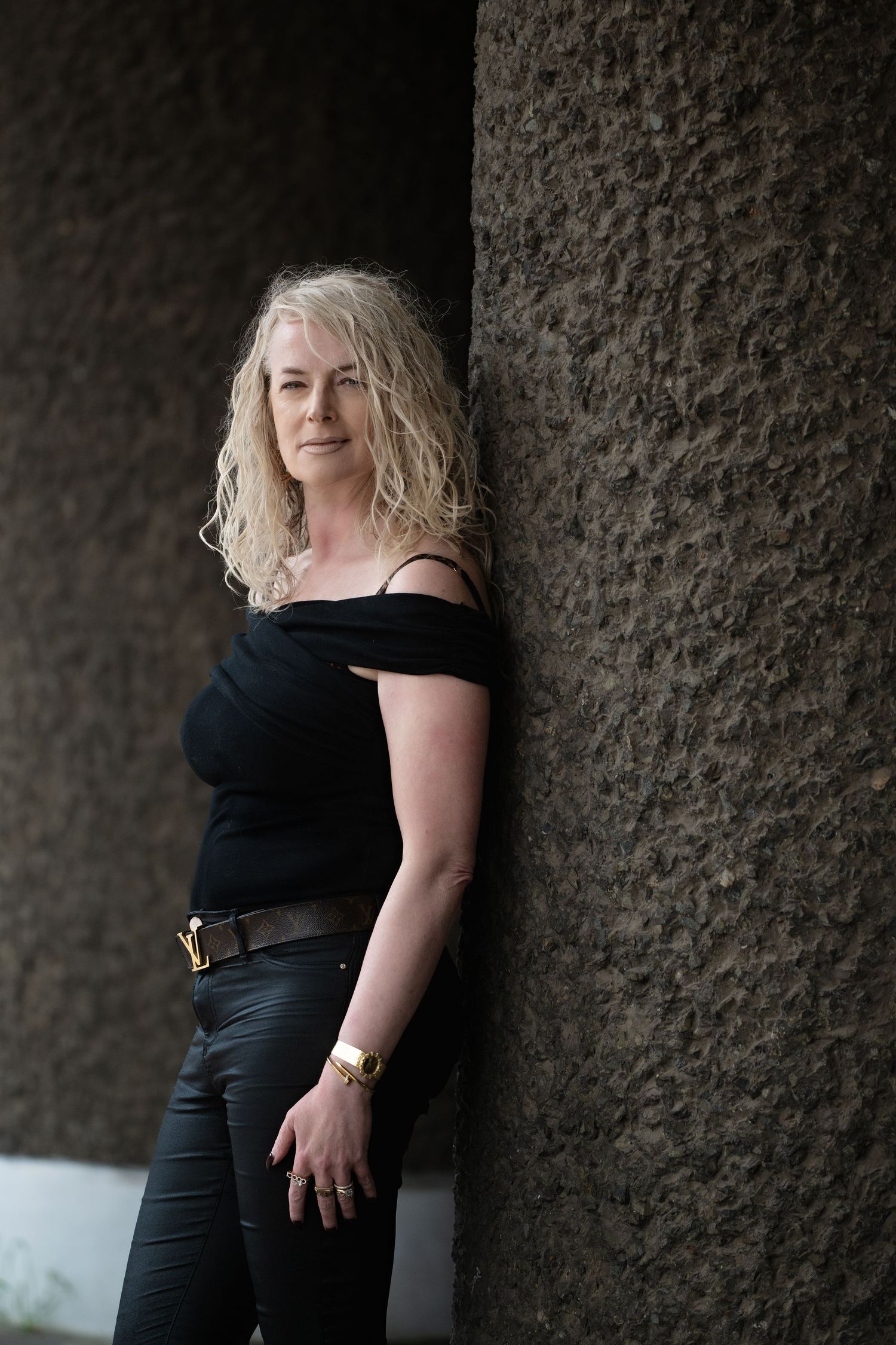 Blonde woman in black off-shoulder top and leather pants leaning against a textured stone wall.