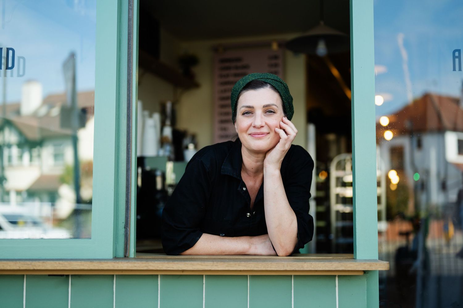 Woman with green hair leaning out of a mint-colored cafe window, smiling warmly with chin resting on hand.