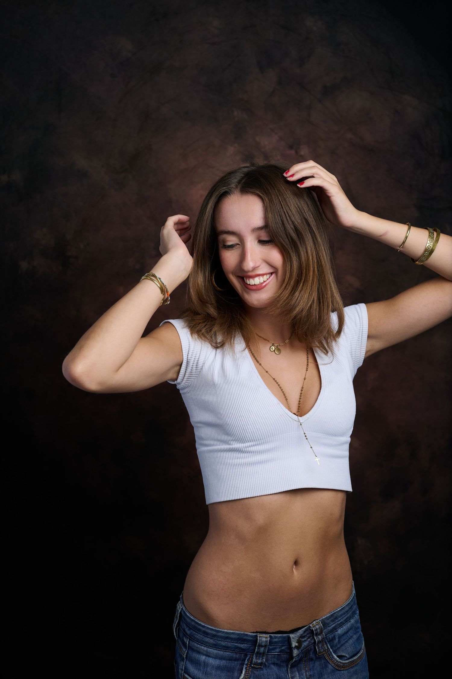 Smiling young woman in white crop top and jeans with hands in hair on dark background.