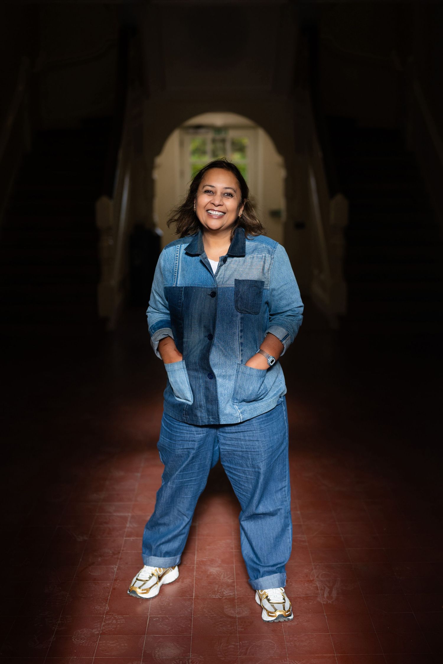 Smiling woman in blue denim outfit standing in a dark hallway with arched doorway.