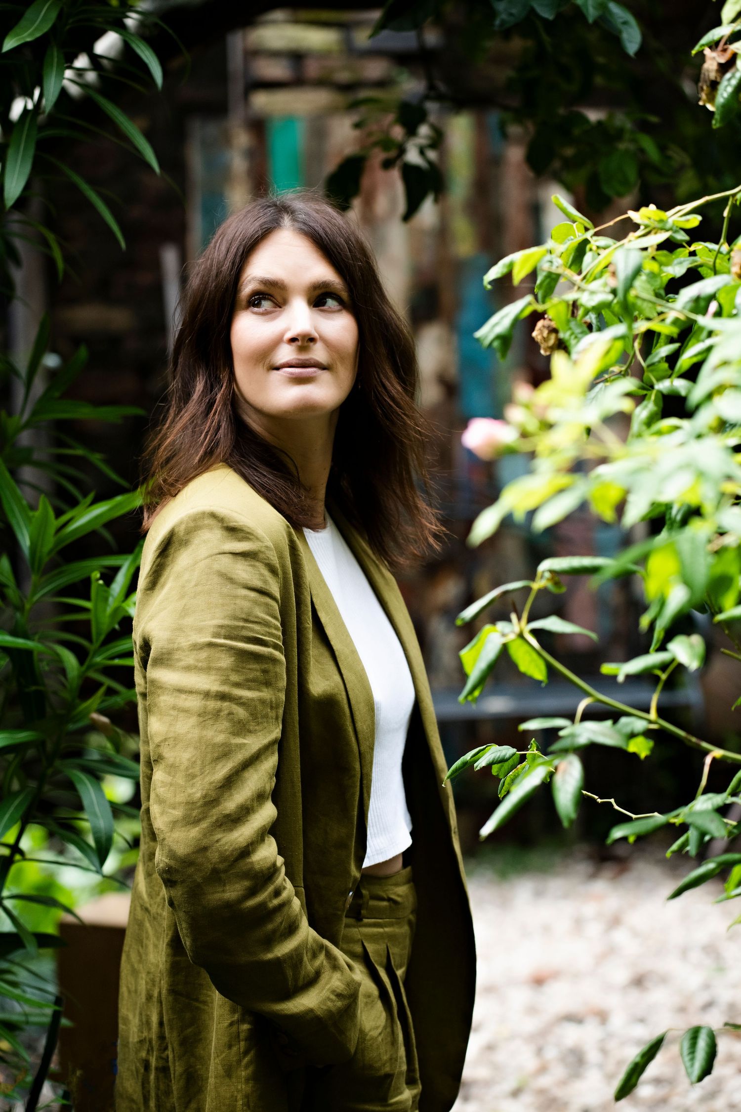 A brunette woman in an olive green jacket stands among lush garden plants, gazing upward in natural light.